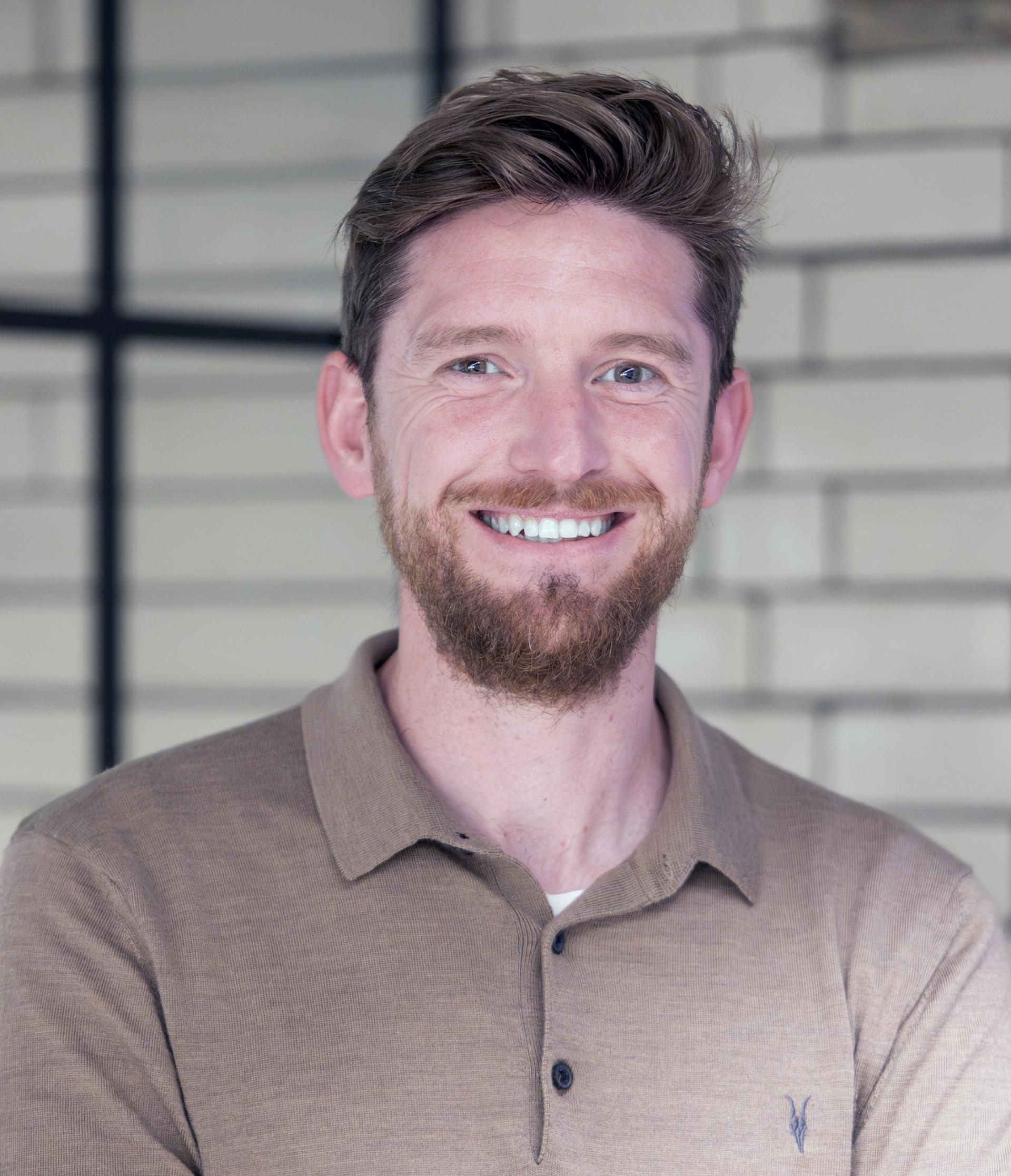 Smiling man with a beard wearing a brown polo shirt, standing against a brick wall, discussing Manchester's residential property outlook.