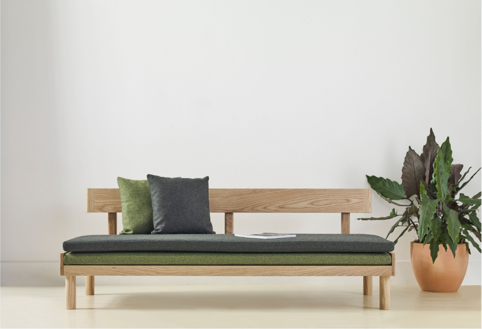 Sleek wooden sofa with green and grey cushions, accompanied by a potted plant, showcasing modern design from the Ori Collection.