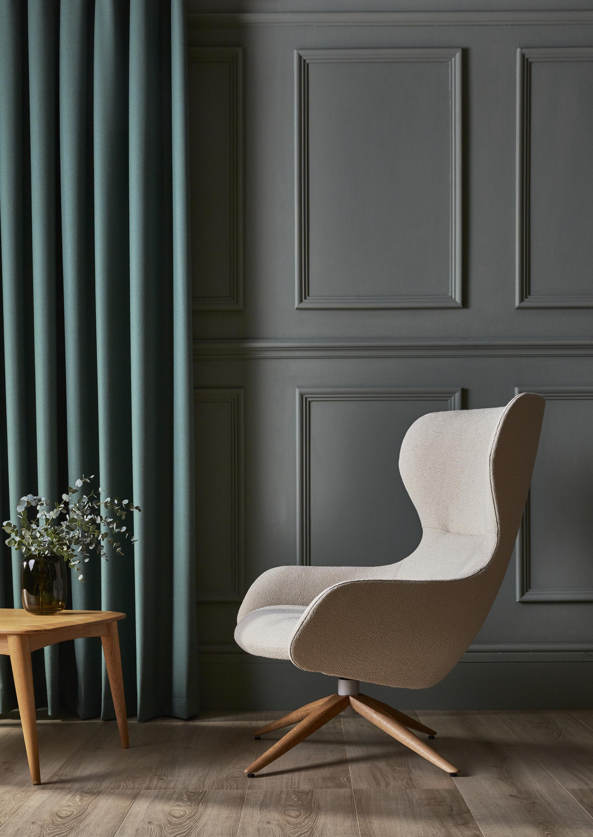 Stylish beige bouclé armchair next to a wooden table with foliage, set against a dark green wall and curtains.