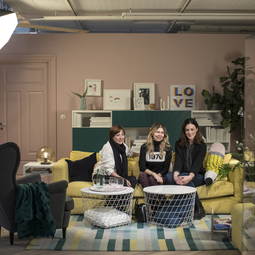 Three women relax on a stylish yellow couch in a modern IKEA-inspired living room with plants and artwork.