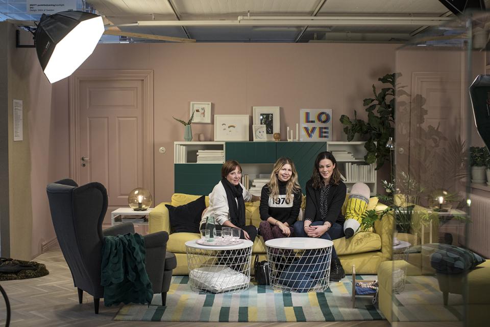 Three women relax on a stylish yellow couch in a modern IKEA-inspired living room with plants and artwork.