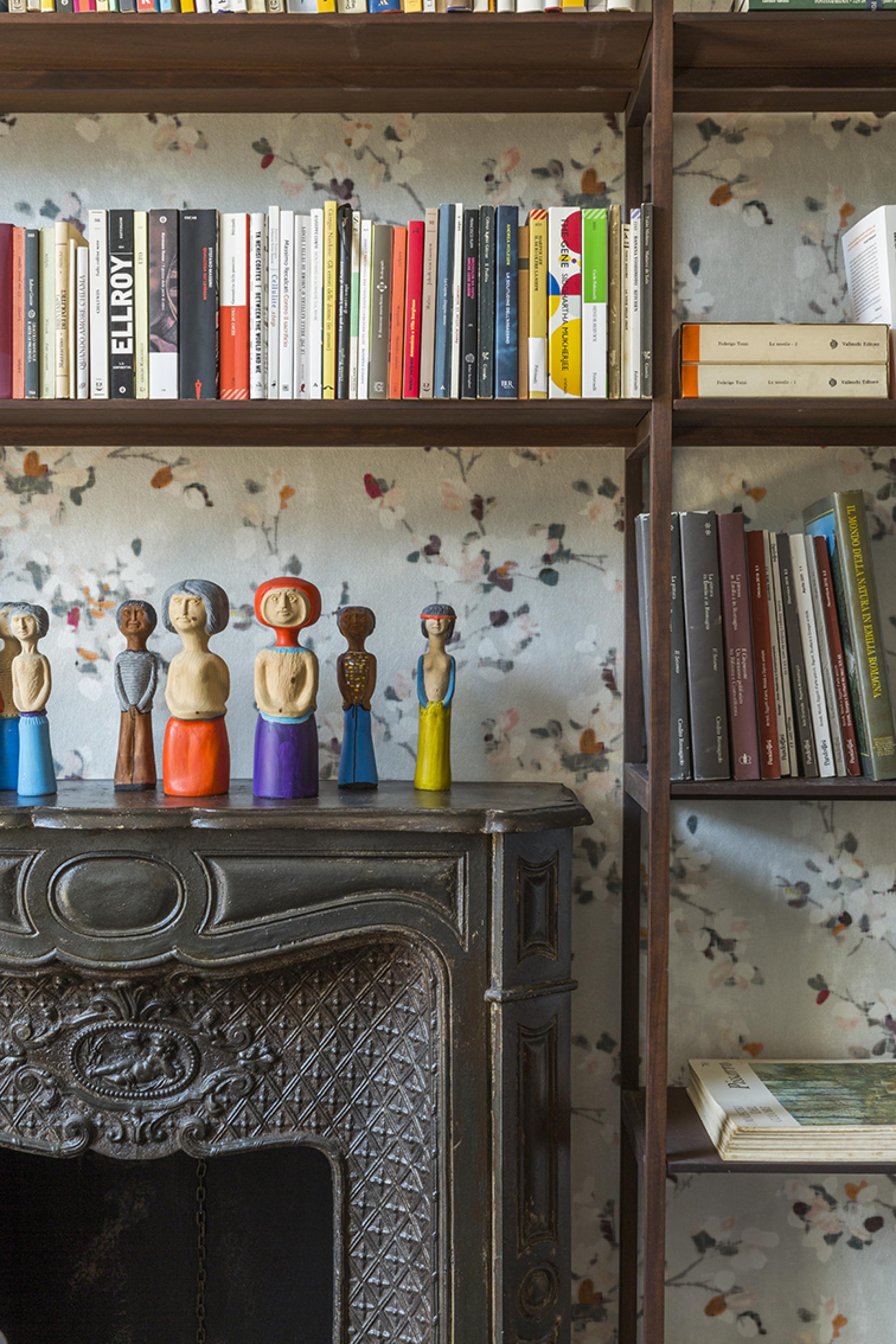 Colorful wooden sculptures atop a vintage fireplace, surrounded by bookshelves filled with diverse literature in an artist's loft.