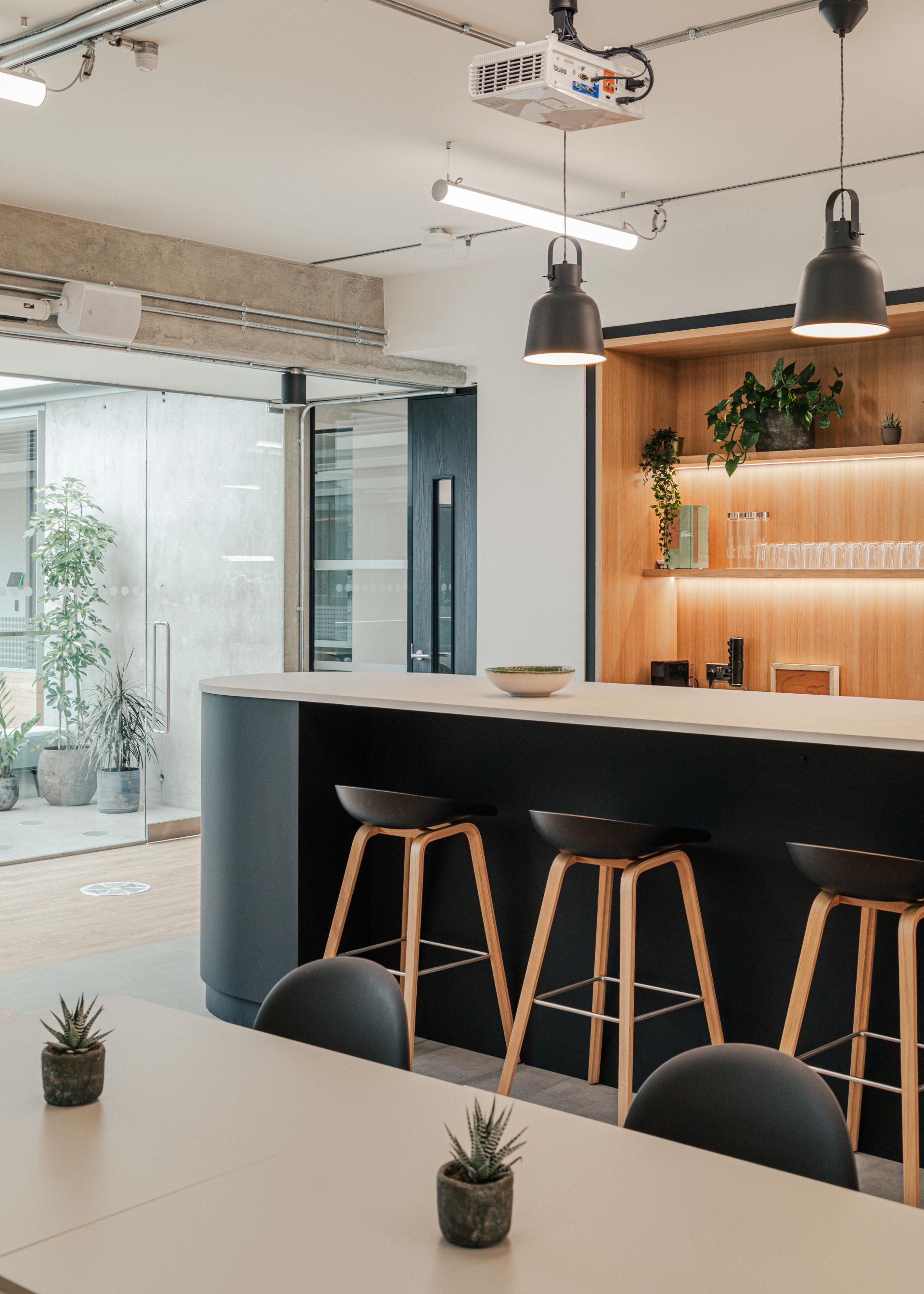 Modern workspace design featuring a sleek bar area with wooden stools, plants, and glass walls, inspired by New York warehouses.