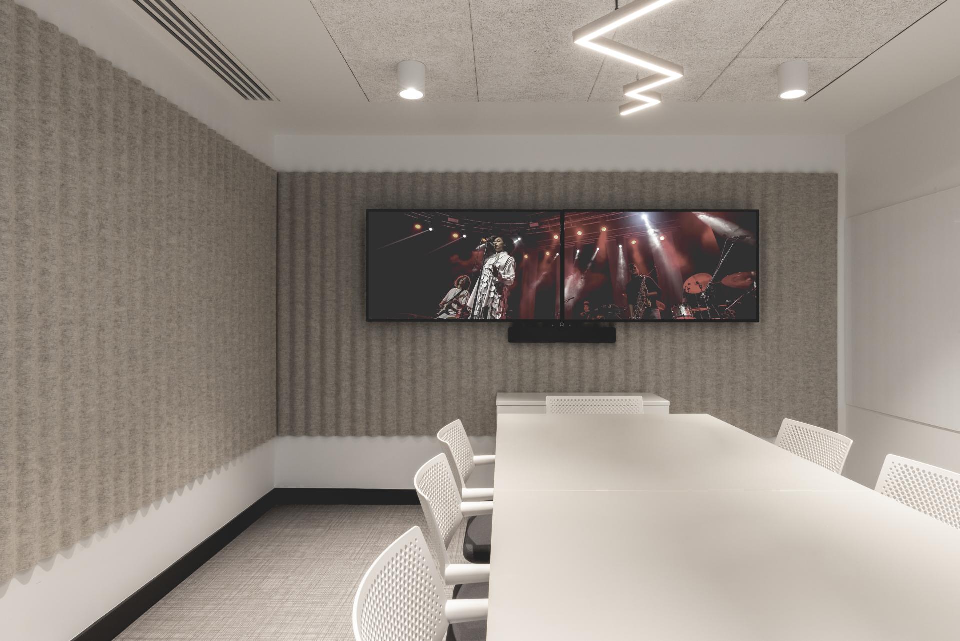 Modern meeting room featuring a minimalist table, white chairs, and dual screens displaying a live music performance.