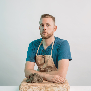 Jeffrey Miller, wearing an apron, poses with clay on a work surface, showcasing his innovative tile project using waste.