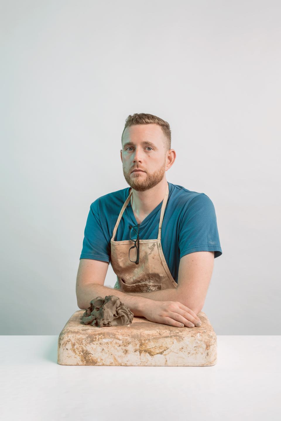 Jeffrey Miller, wearing an apron, poses with clay on a work surface, showcasing his innovative tile project using waste.