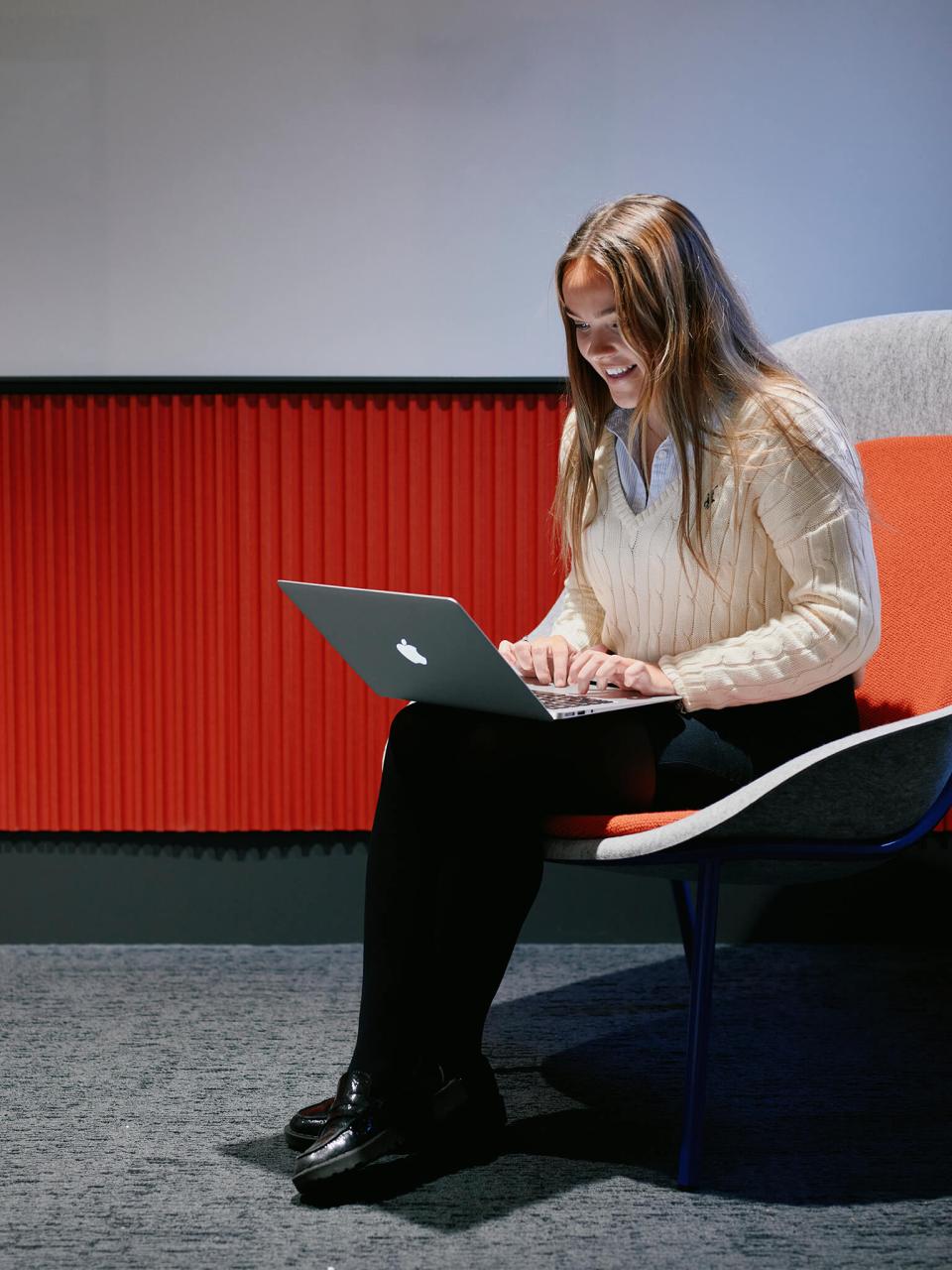 A student sits on a modern chair, focused on her laptop, with vibrant red walls enhancing the study space.