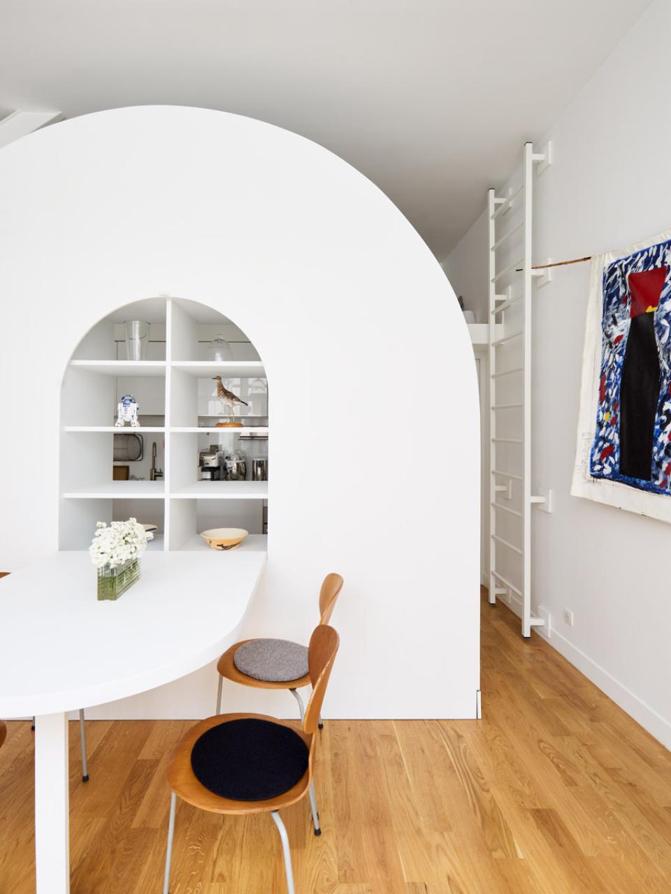 Stylish Parisian living space featuring curved white architecture, minimalist dining table, and wooden chairs with unique cushions.