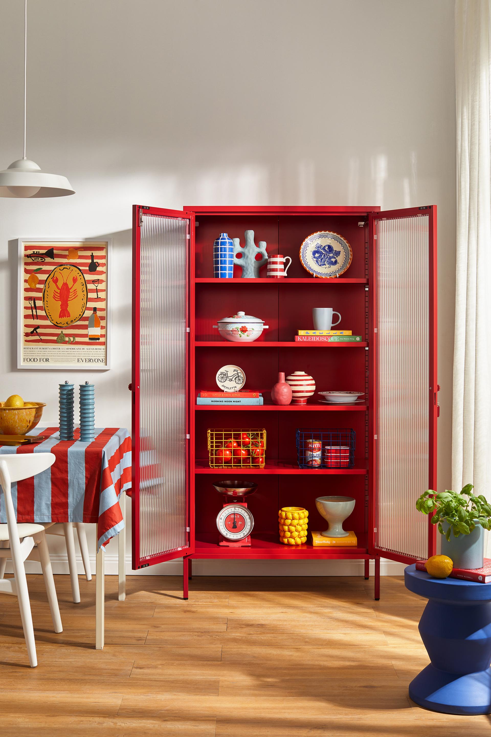 Bright red cabinet displaying Mustard Made's Glass Collection, filled with colorful tableware, decorative items, and vibrant home decor.