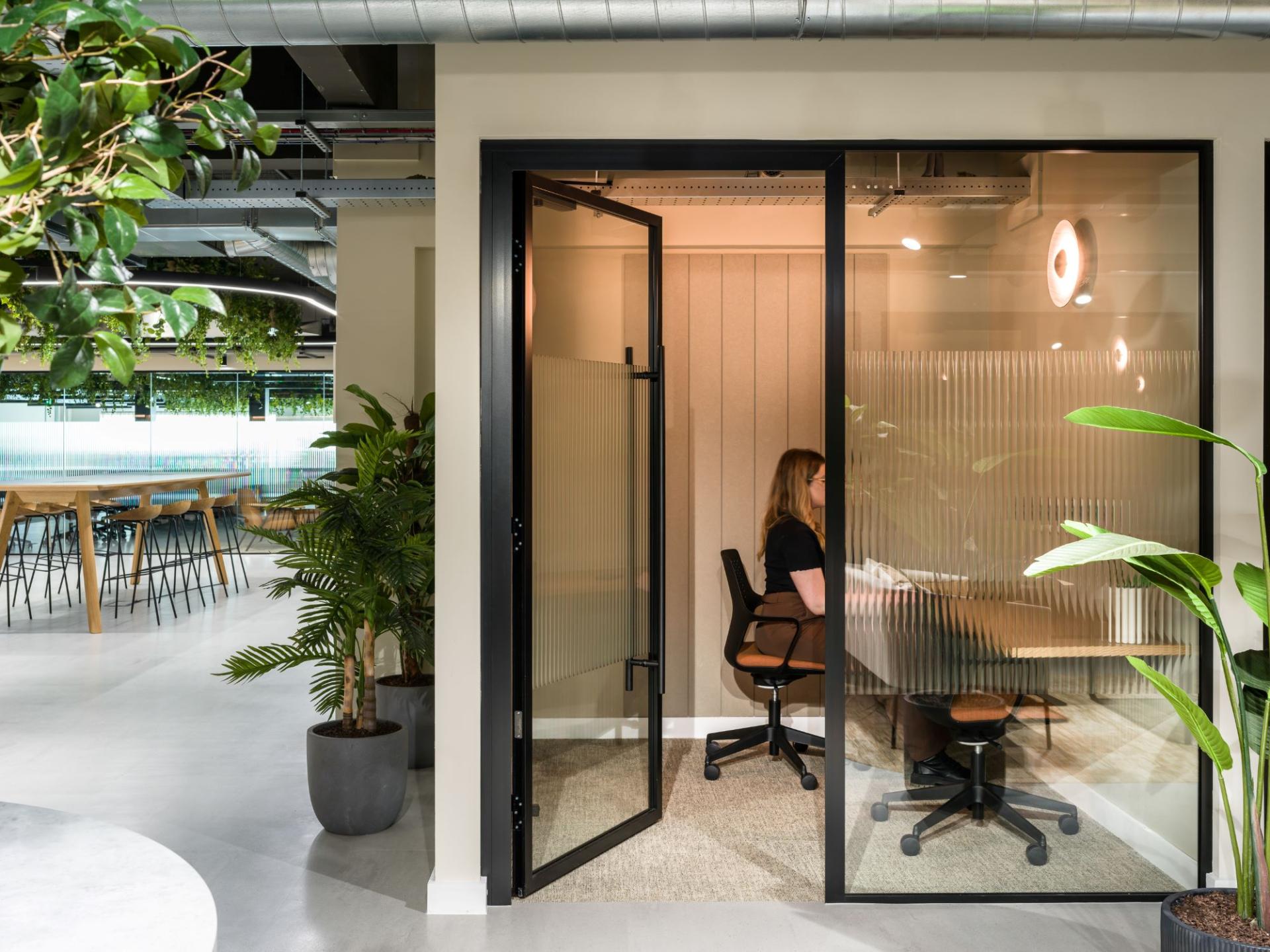 Modern office space at Alderley Park features a glass meeting pod with a person seated and surrounded by greenery.