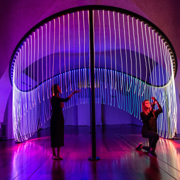 Interactive light installation at London Design Biennale featuring colorful, flowing LED strands and engaged visitors.
