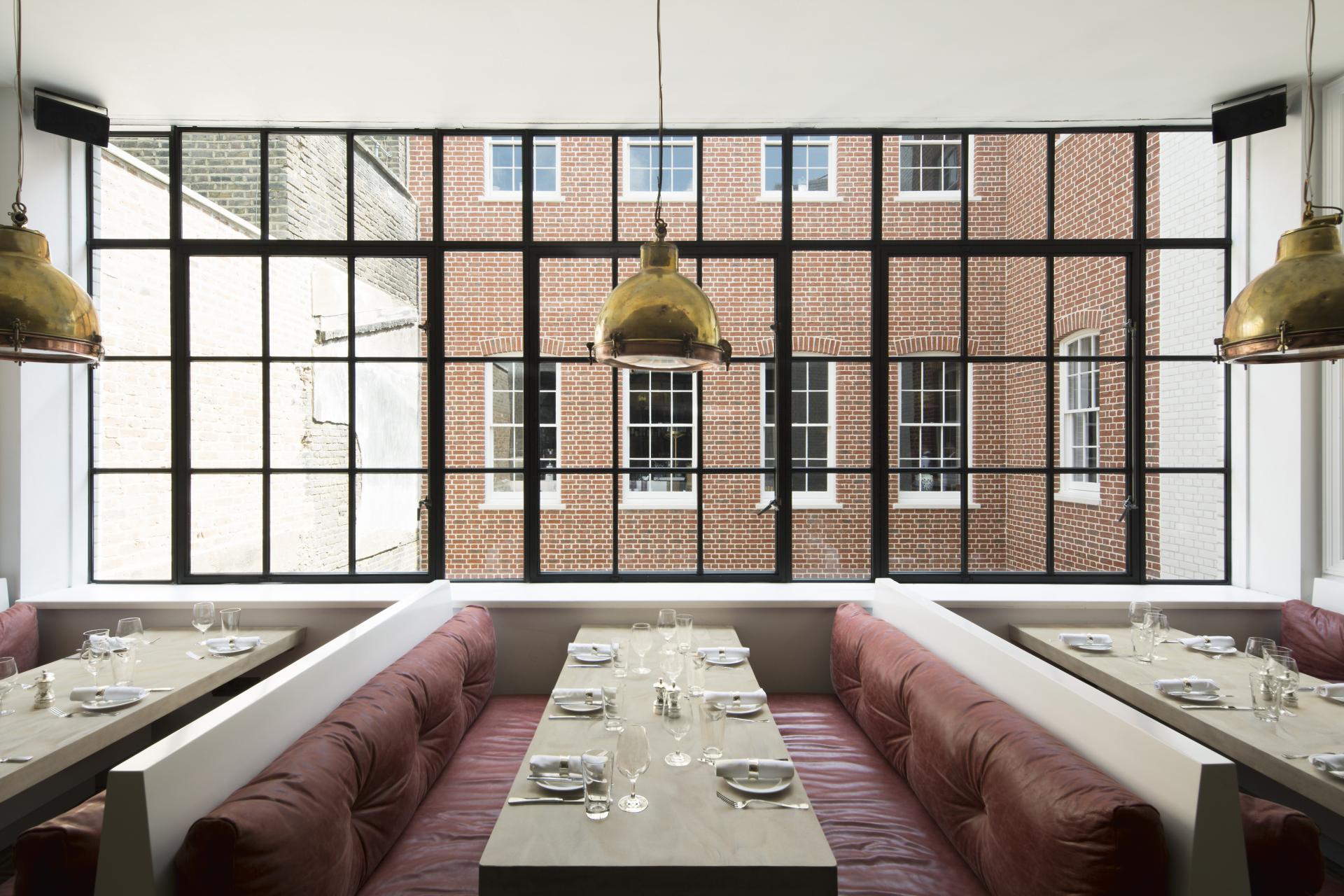 Modern restaurant interior featuring leather seating, stone tables, and large windows with a view of brick buildings.