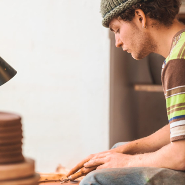A young artisan crafts pottery, focused on his work, surrounded by tools and raw materials in a studio setting.