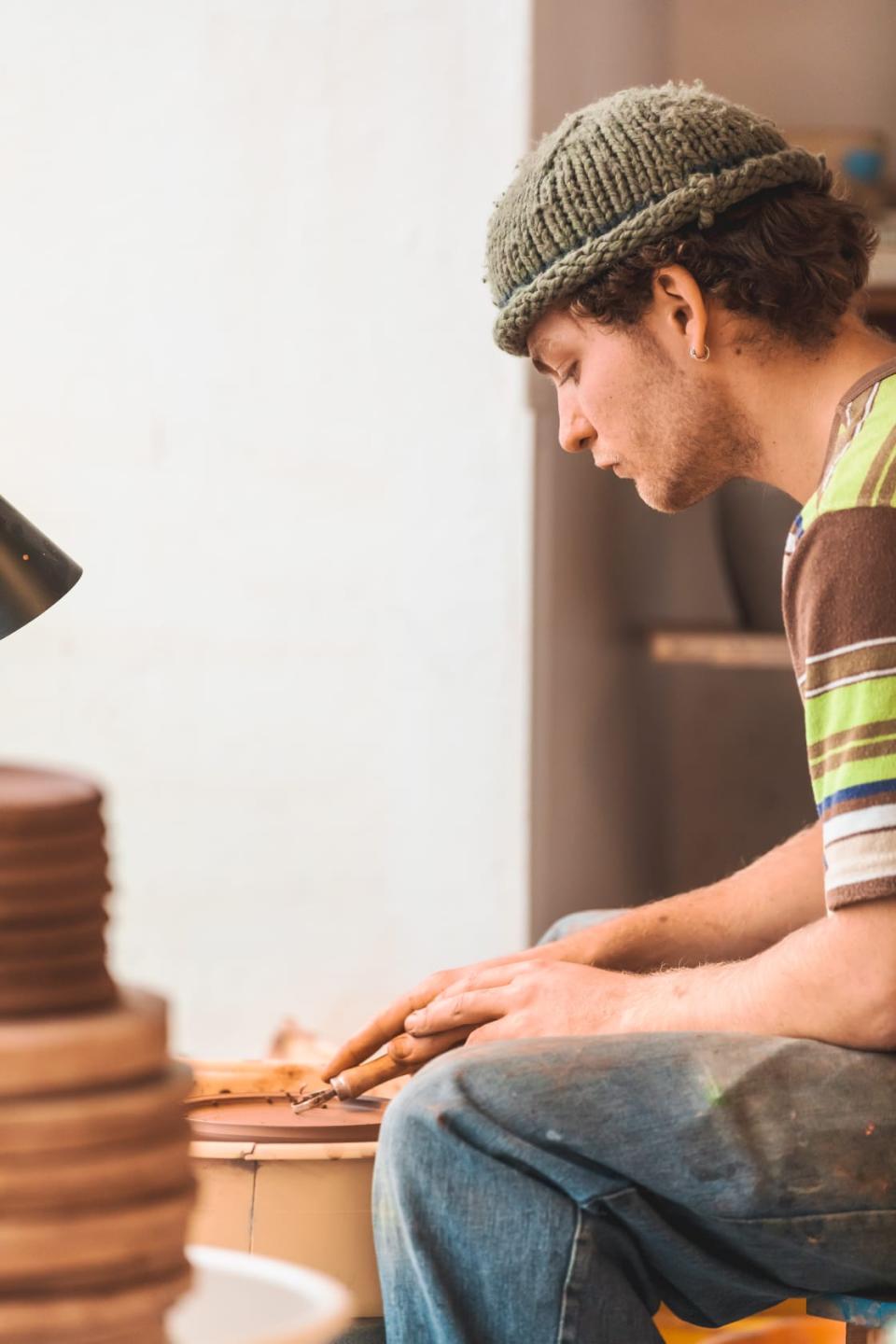 A young artisan crafts pottery, focused on his work, surrounded by tools and raw materials in a studio setting.