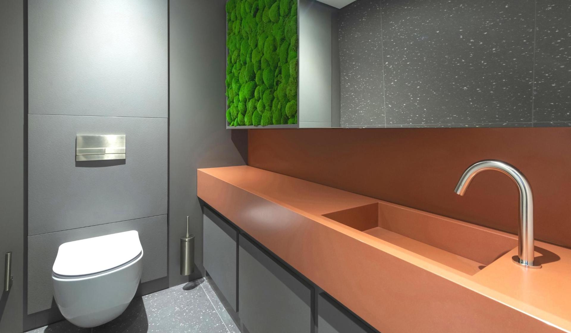 Modern commercial washroom design featuring a sleek orange countertop, minimalist sink, and vibrant green moss wall for wellness.