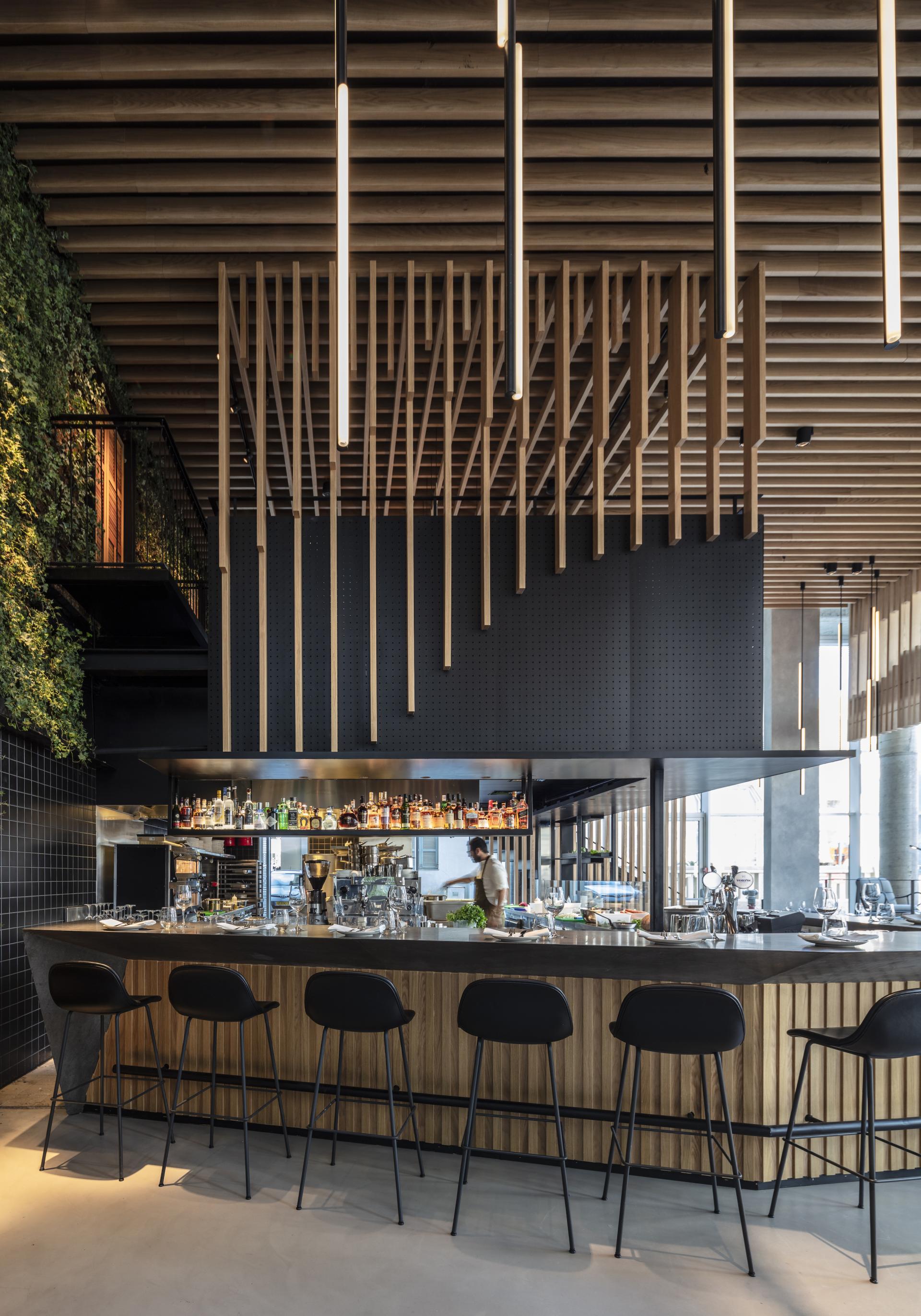 Sleek bar area in Tel Aviv restaurant featuring suspended wooden beams and lush greenery, designed by Kimmel Eshkolot Architects.