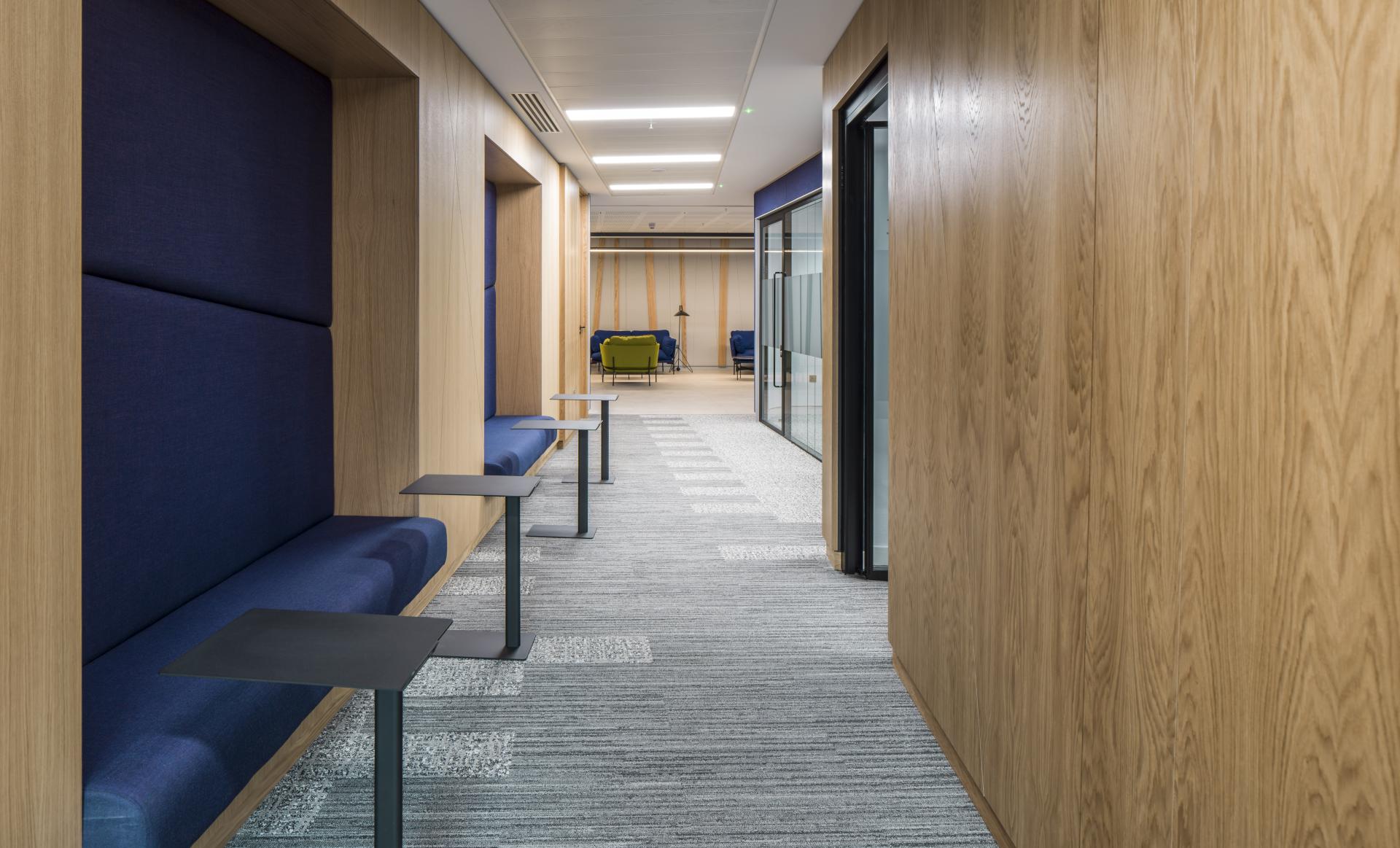 Modern office hallway featuring wooden walls, blue seating areas, and collaborative workspaces, showcasing the WELL certified design by Form Design Consultants.
