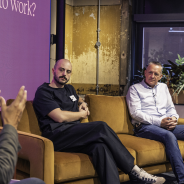 Two speakers engaged in discussion on technology's role in the modern workplace, with a backdrop displaying a relevant question.