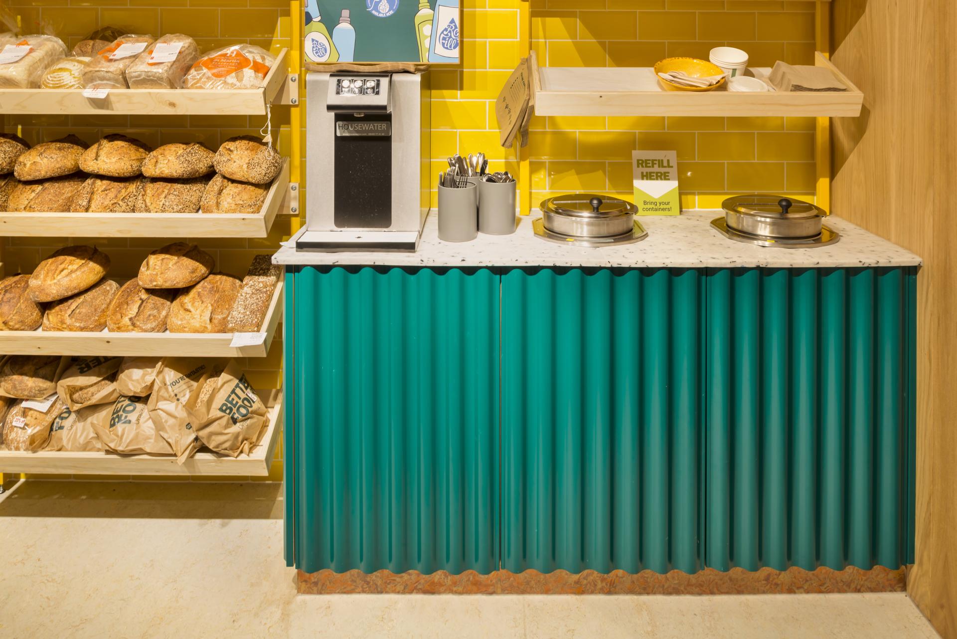 Brightly colored café-deli counter with bread displays, refill station, and utensils, designed for Better Food's new Gloucester Road store.