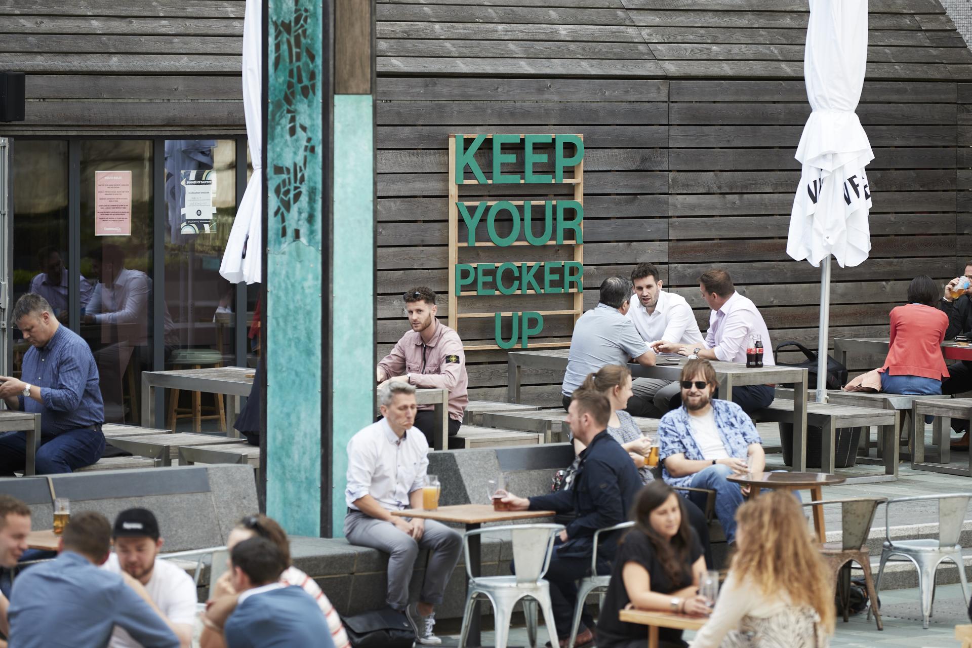 Colourful outdoor sign reading "KEEP YOUR PECKER UP" amid people enjoying drinks in Manchester's NOMA neighbourhood.