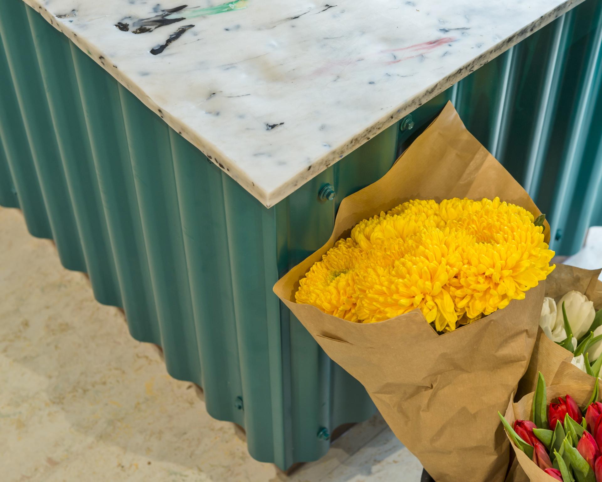 Bright yellow chrysanthemums in brown paper wrapping, displayed next to a marble-topped green counter in a café-deli setting.