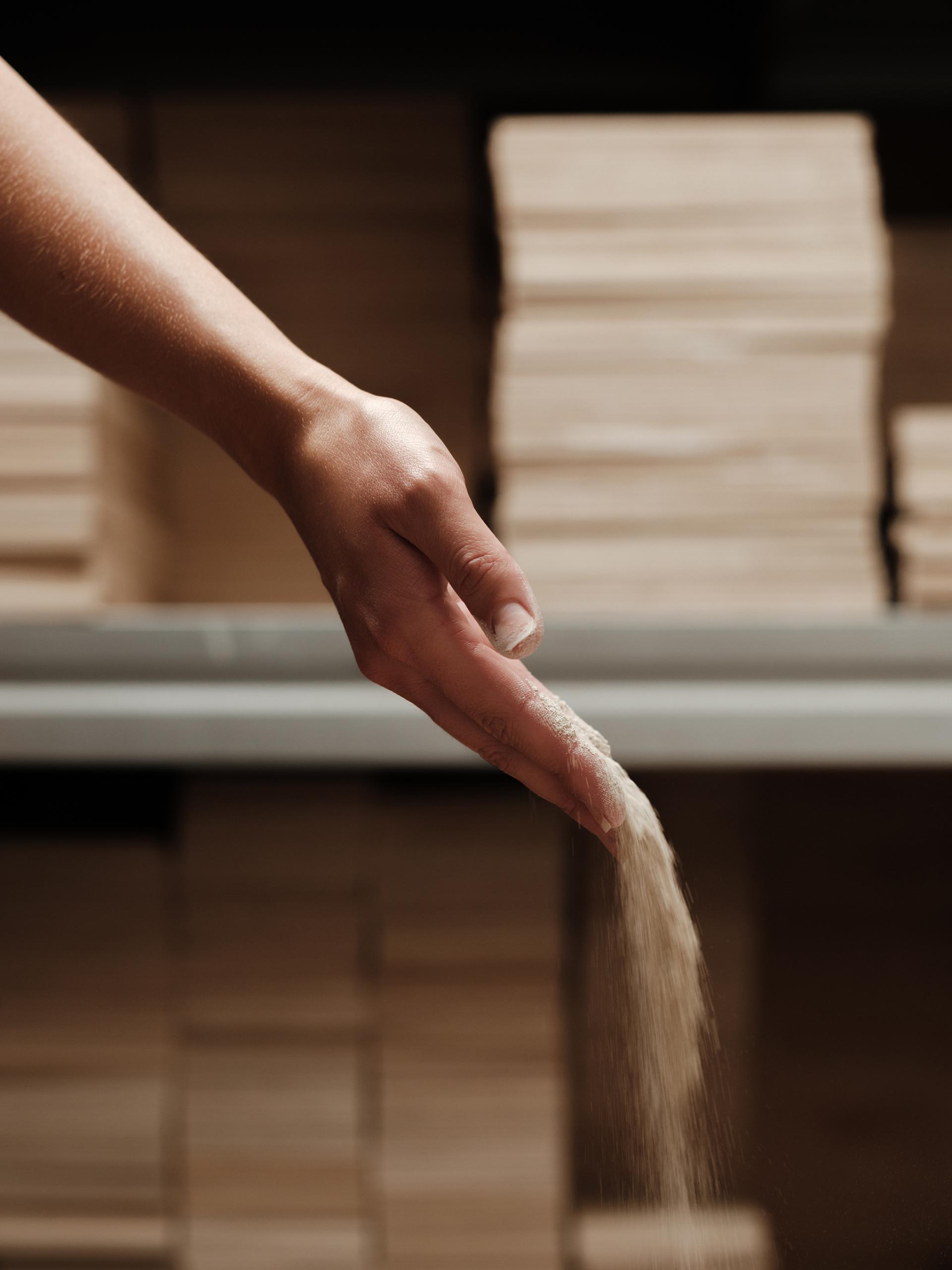 Hand pouring fine material over stacked wooden boards, highlighting sustainable practices in clay and wall finishes production.