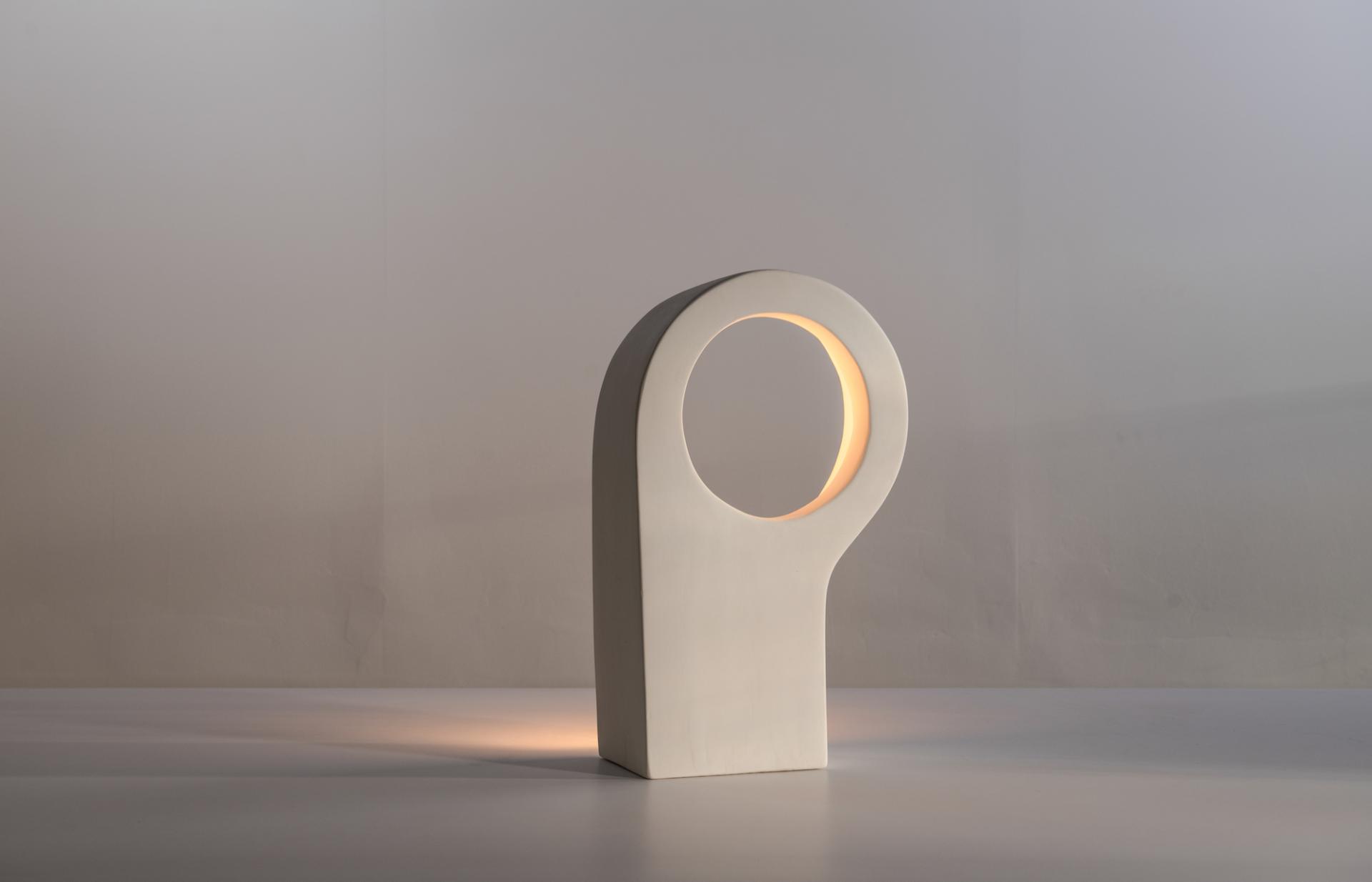 Sculptural artwork featuring a modern white lamp with an oval cutout, illuminated warmly, showcased at Reynold Rodriguez's solo exhibition.
