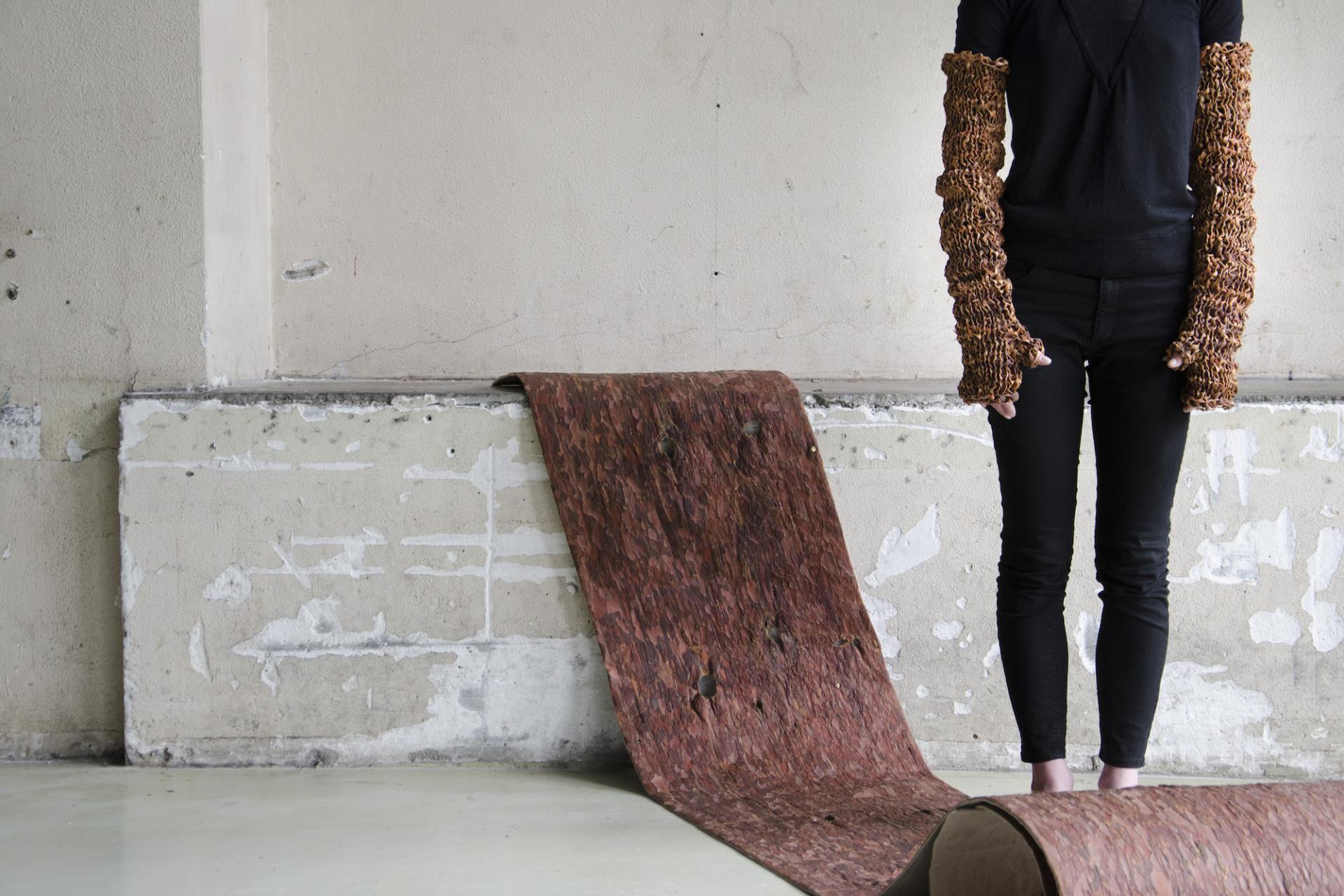 A person stands in a minimalist space wearing innovative biomaterials arm warmers, next to a rolled biomaterials textile.