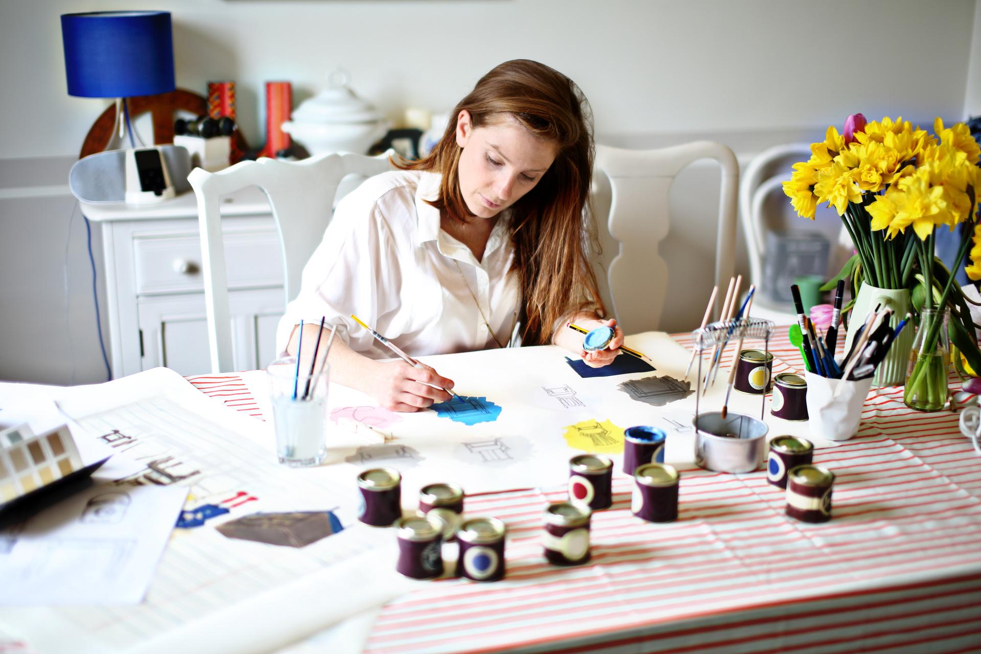 A woman paints design sketches at a table, surrounded by paint pots, brushes, and flowers, showcasing creativity in interior design.