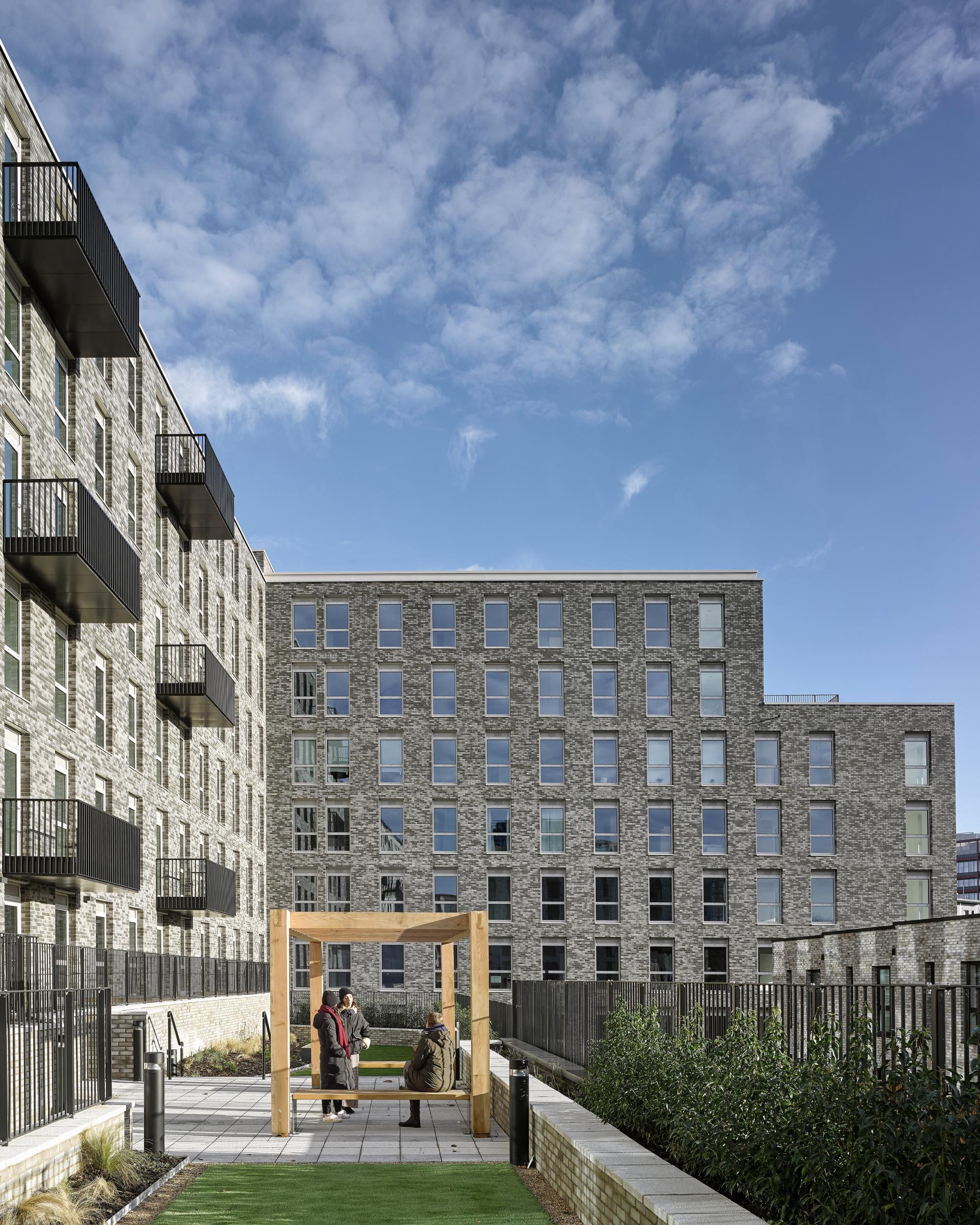 Urban outdoor space featuring three individuals conversing beneath a wooden structure, surrounded by modern residential architecture and greenery.
