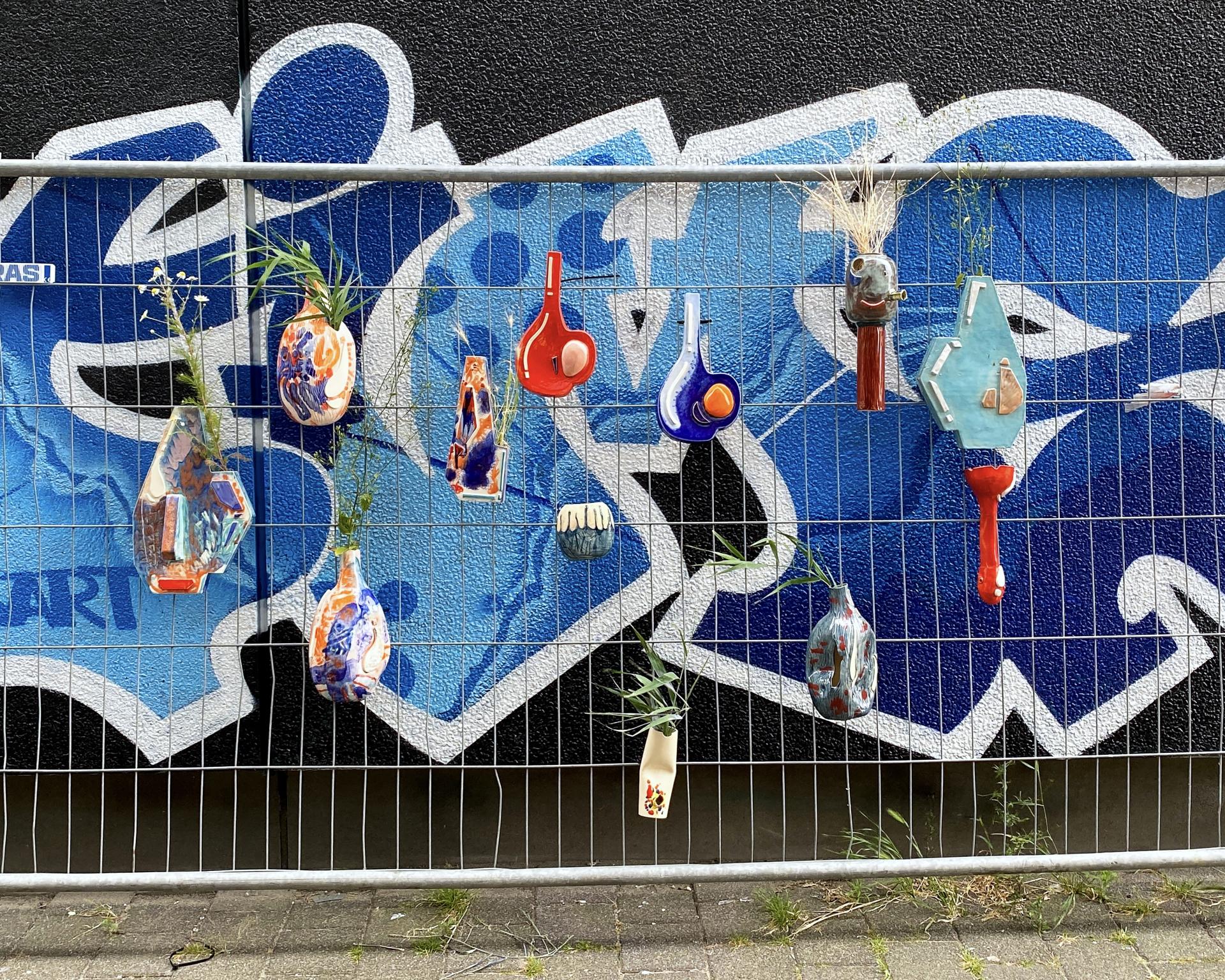 Ceramic vases adorned with vibrant colors, hanging on a fence in front of a blue graffiti backdrop.