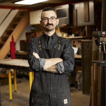 Brooklyn-based maker stands confidently in his workshop, showcasing craftsmanship and creativity in design.