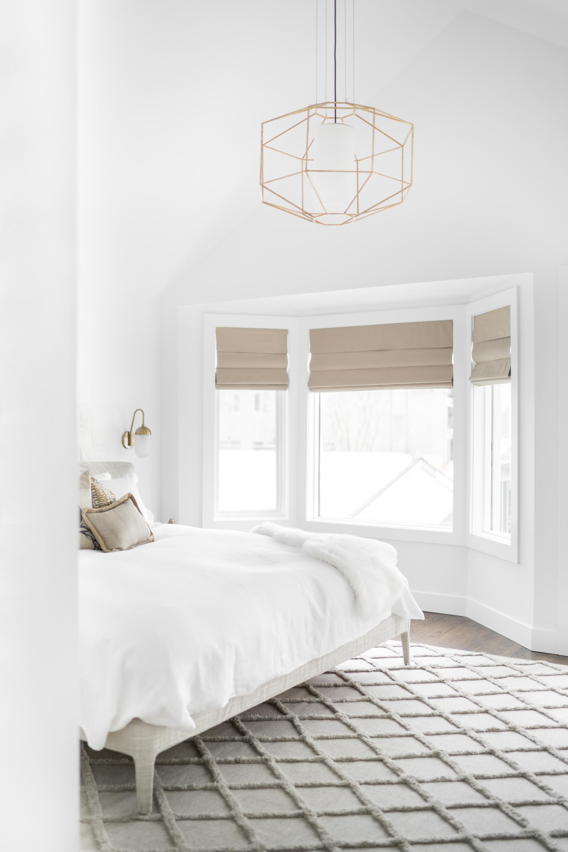 Stylish modern chandelier hangs above a serene bedroom with a cozy bed and large windows.