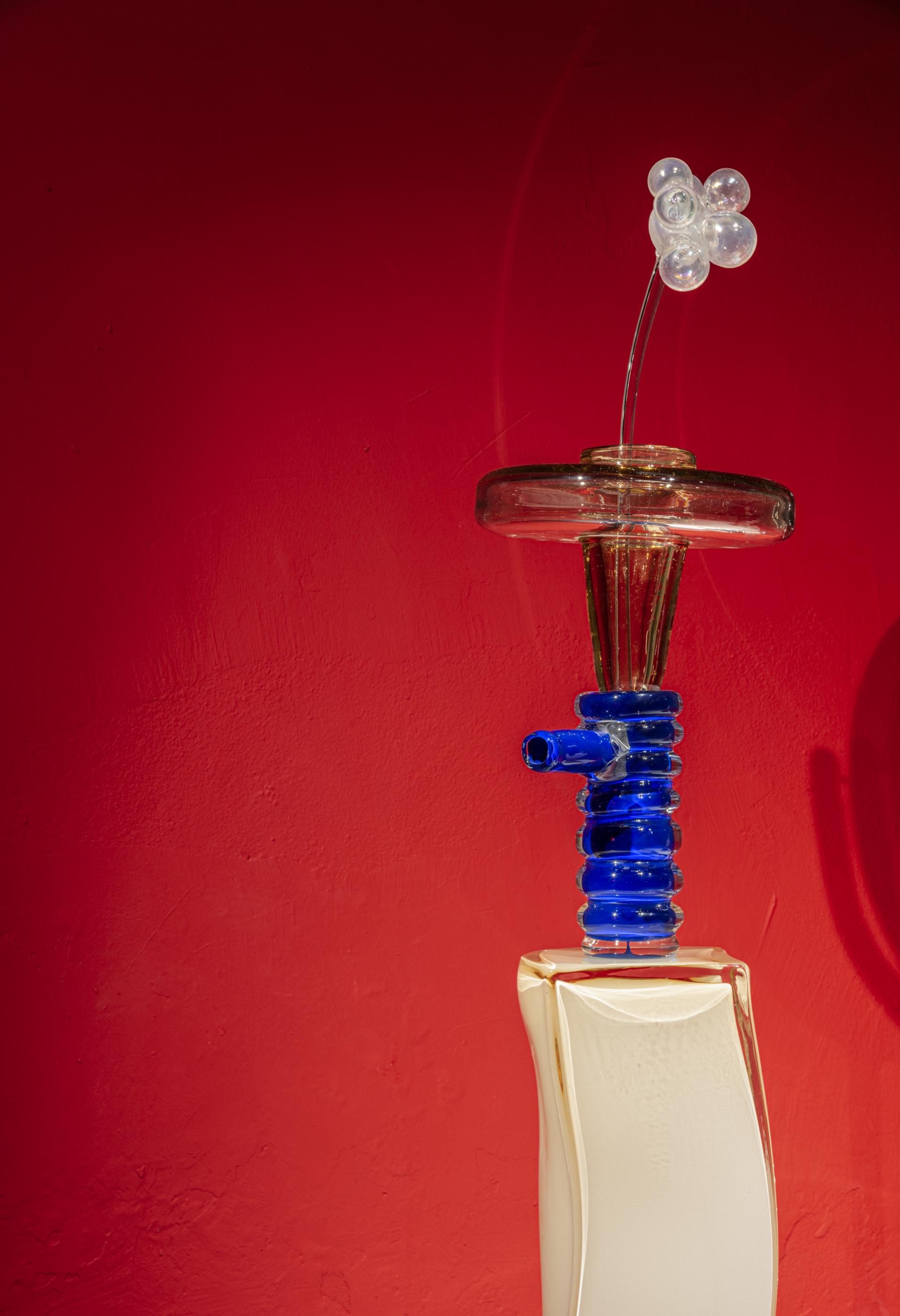 Colorful glass sculpture featuring a white and blue design, displayed against a vibrant red background at the GLASS to GLASS exhibit.