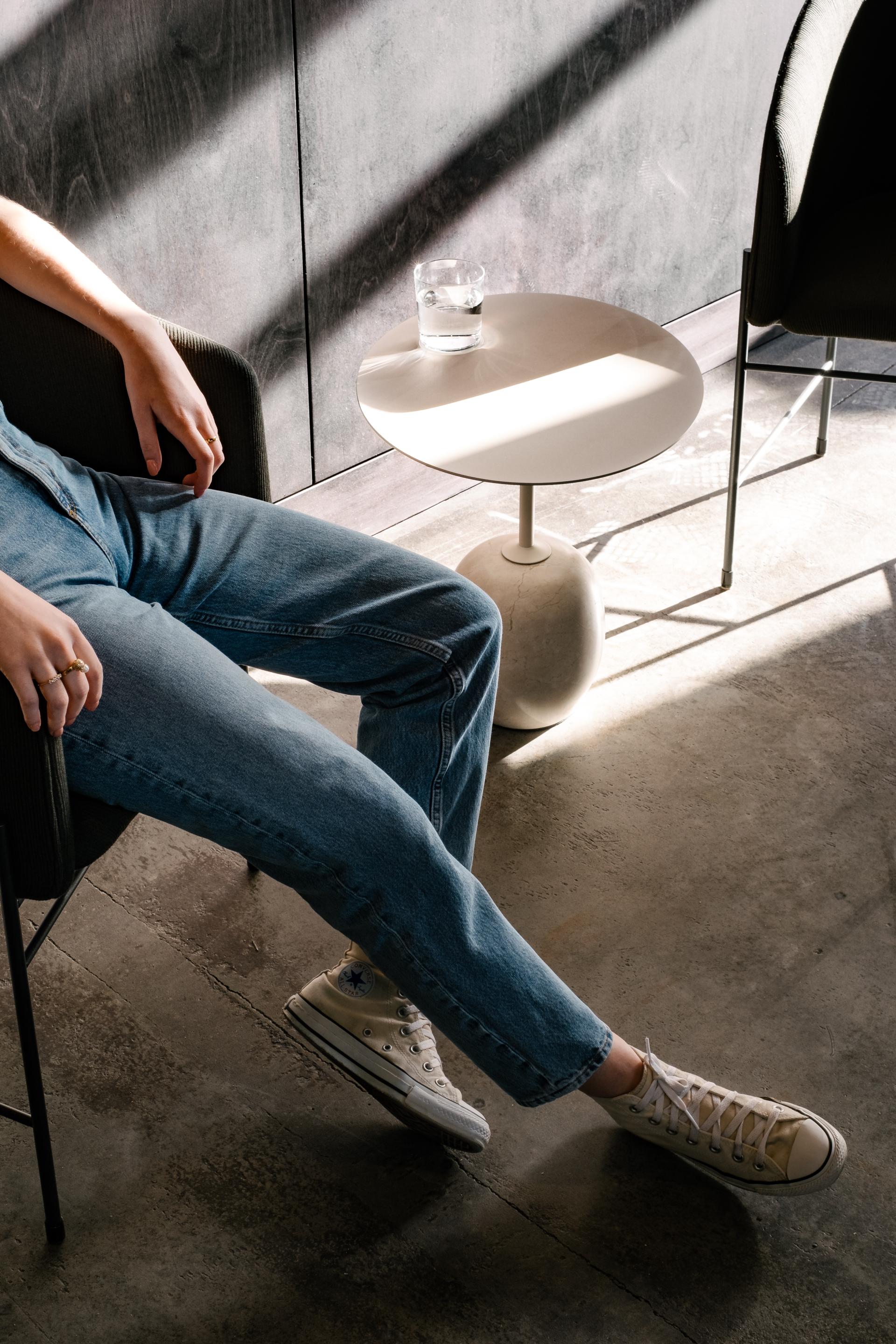 Casual workspace setting featuring a person in denim jeans and sneakers next to a minimalist side table with a glass of water.
