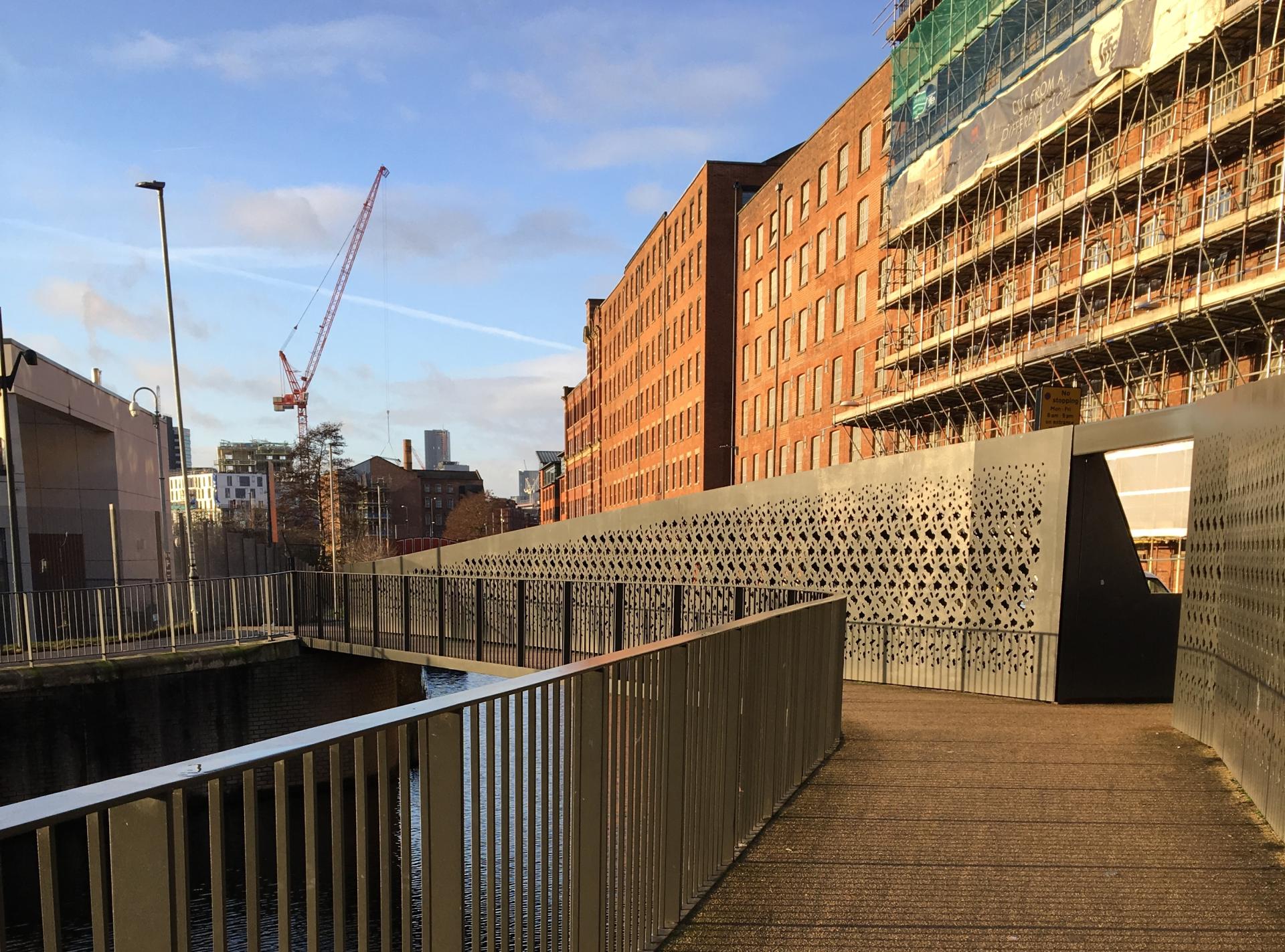Contemporary pedestrian bridge alongside Manchester's historic architecture and construction site, showcasing urban regeneration and design.