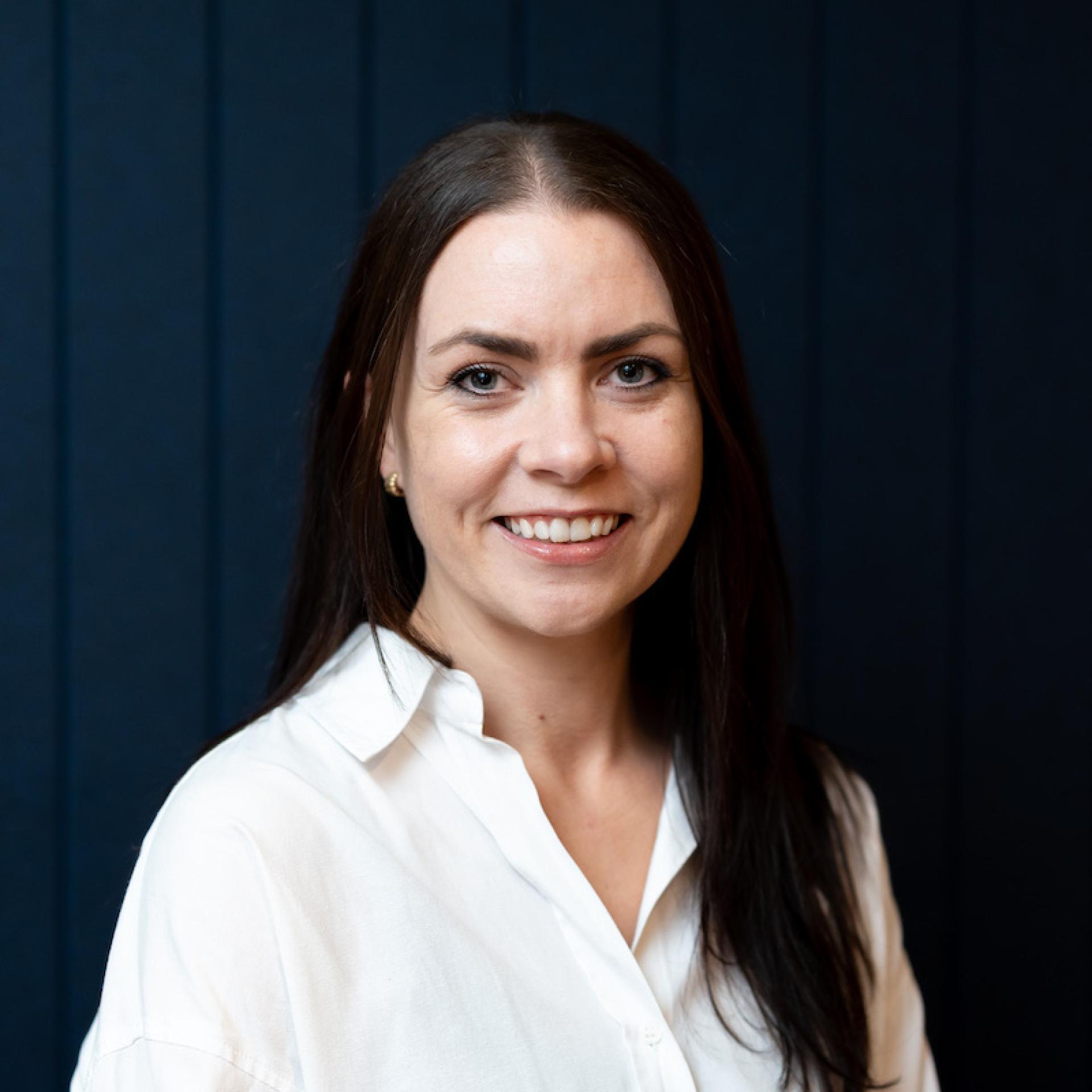 Professional woman in a white shirt smiling, against a navy blue backdrop, reflecting modern workplace dynamics.