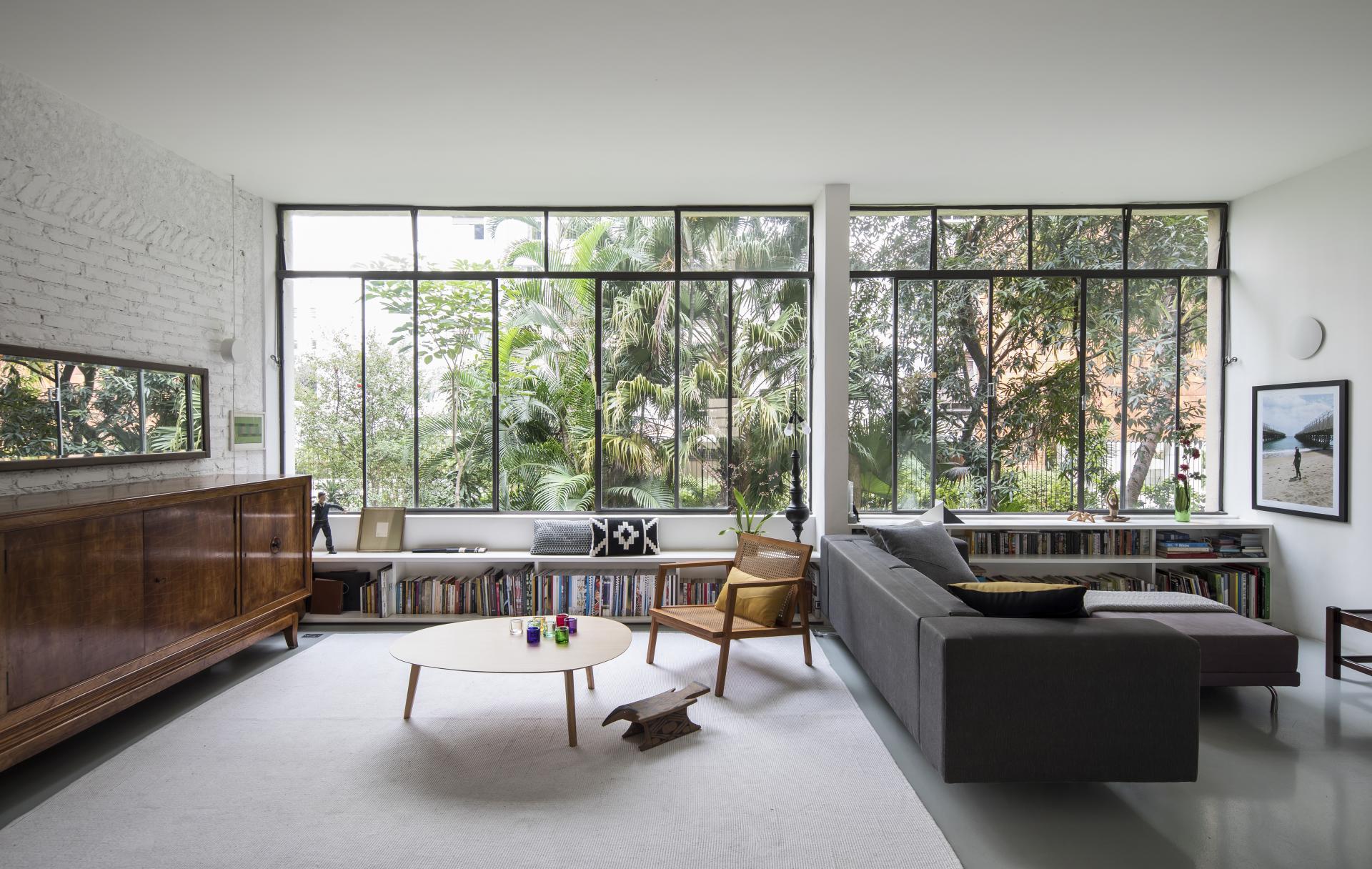 Modern mid-century living room in São Paulo featuring large windows, greenery, and stylish furnishings by Cupertino Arquitetura.