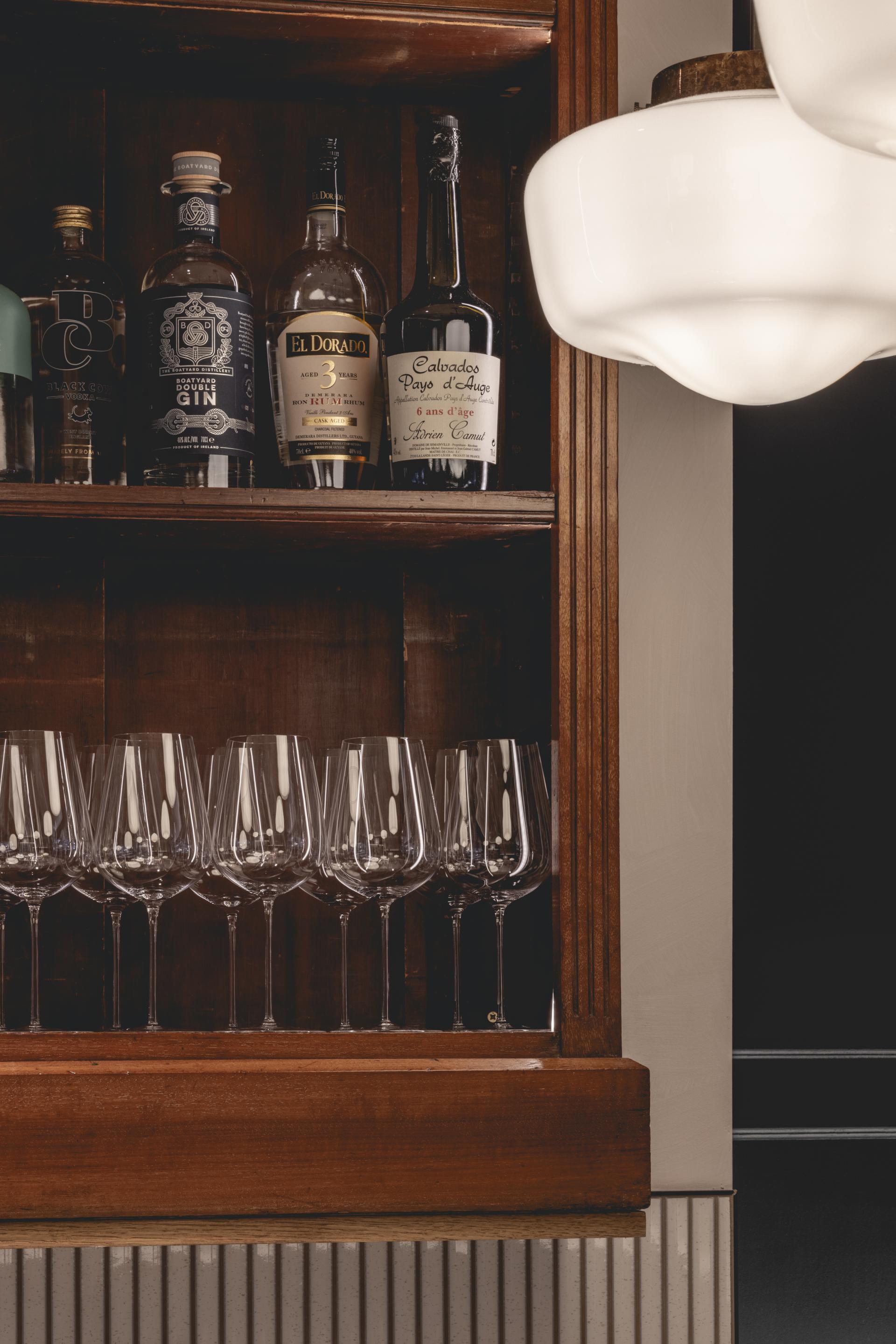 Elegant display of spirits and wine glasses in a vintage wooden cabinet, reflecting Parisian bistro and British countryside design.