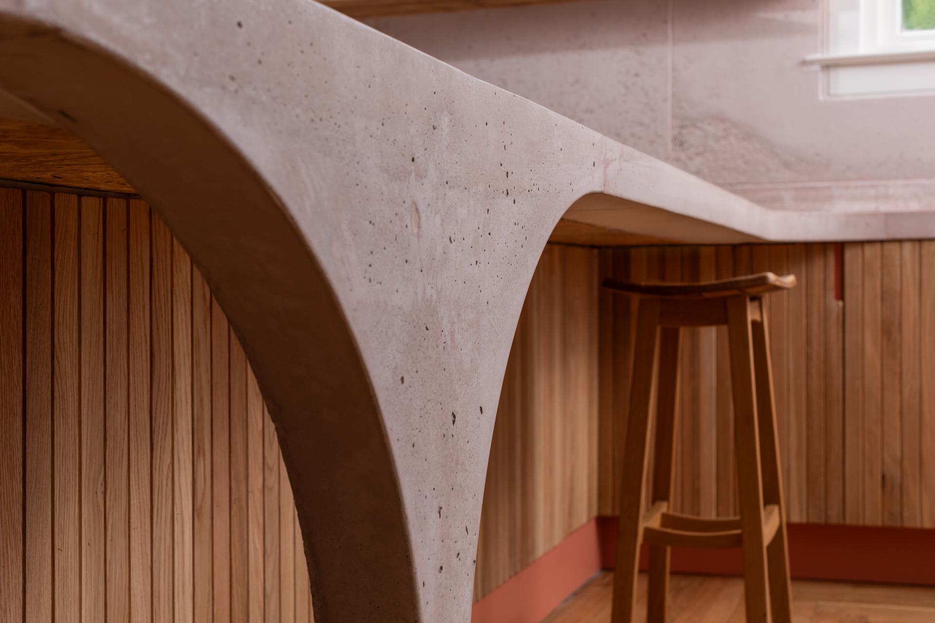 Curved pink concrete countertop and Scottish Oak paneling create a modern, inviting kitchen space with a wooden stool.