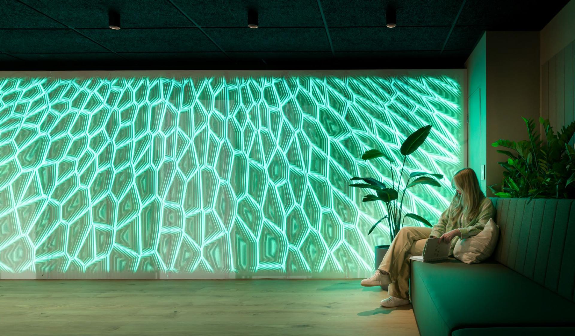 Modern workspace featuring a green illuminated wall design, plants, and a person reading while relaxing on a couch.