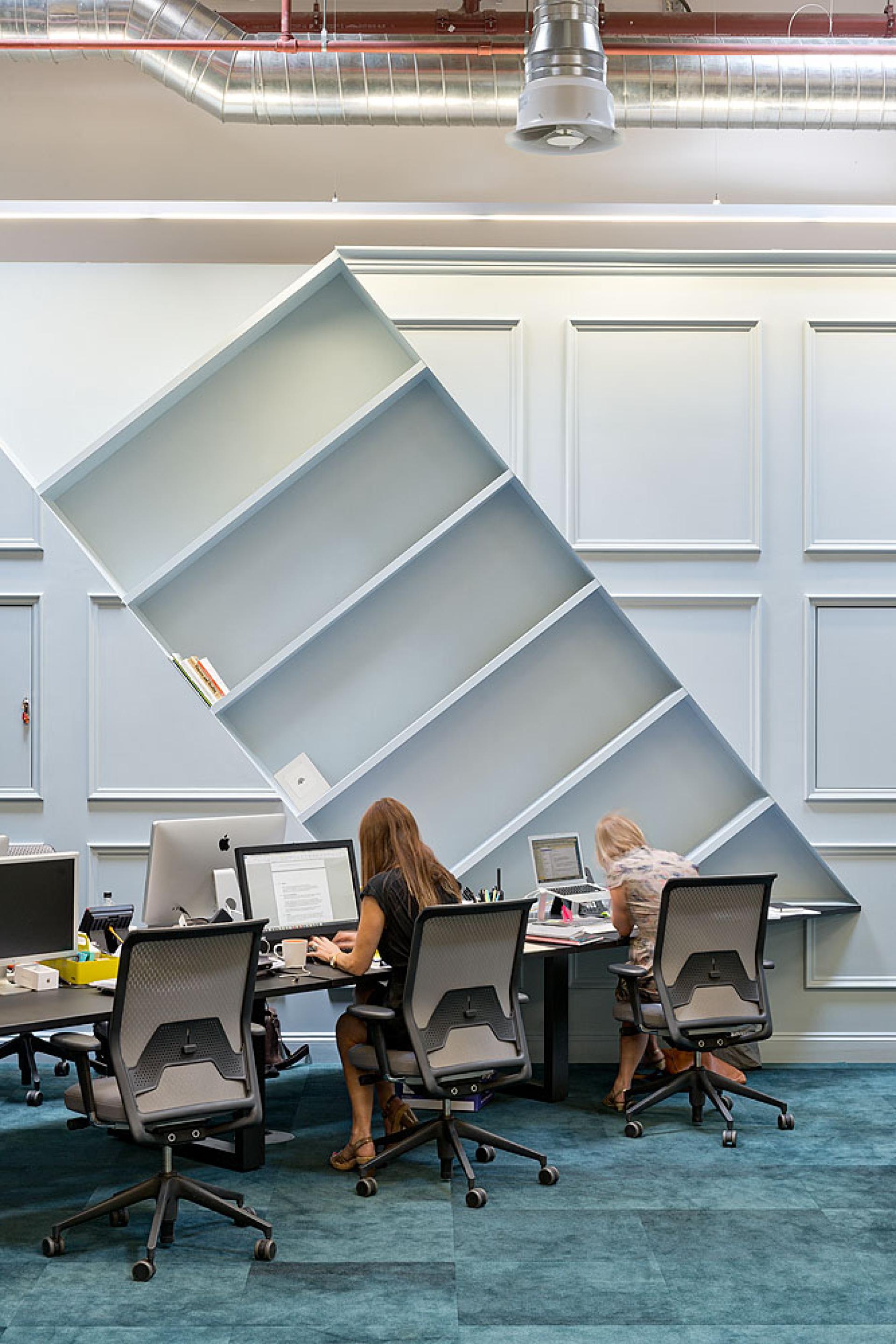 Modern office interior featuring a playful, tilted bookshelf and designers collaborating at workstations, highlighting innovative interior architecture.