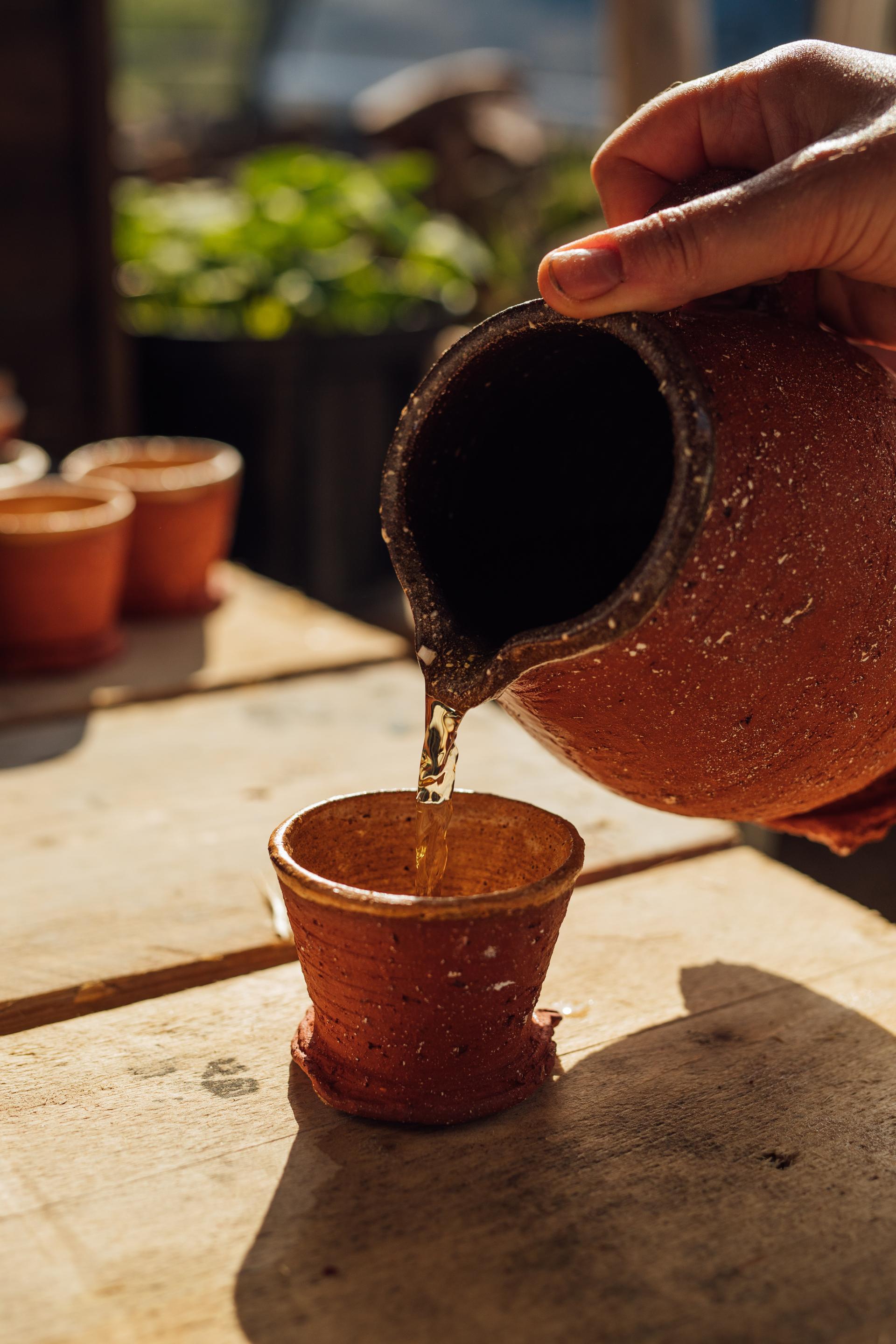 Hand pouring liquid from a clay pitcher into a small terracotta cup on a wooden surface.
