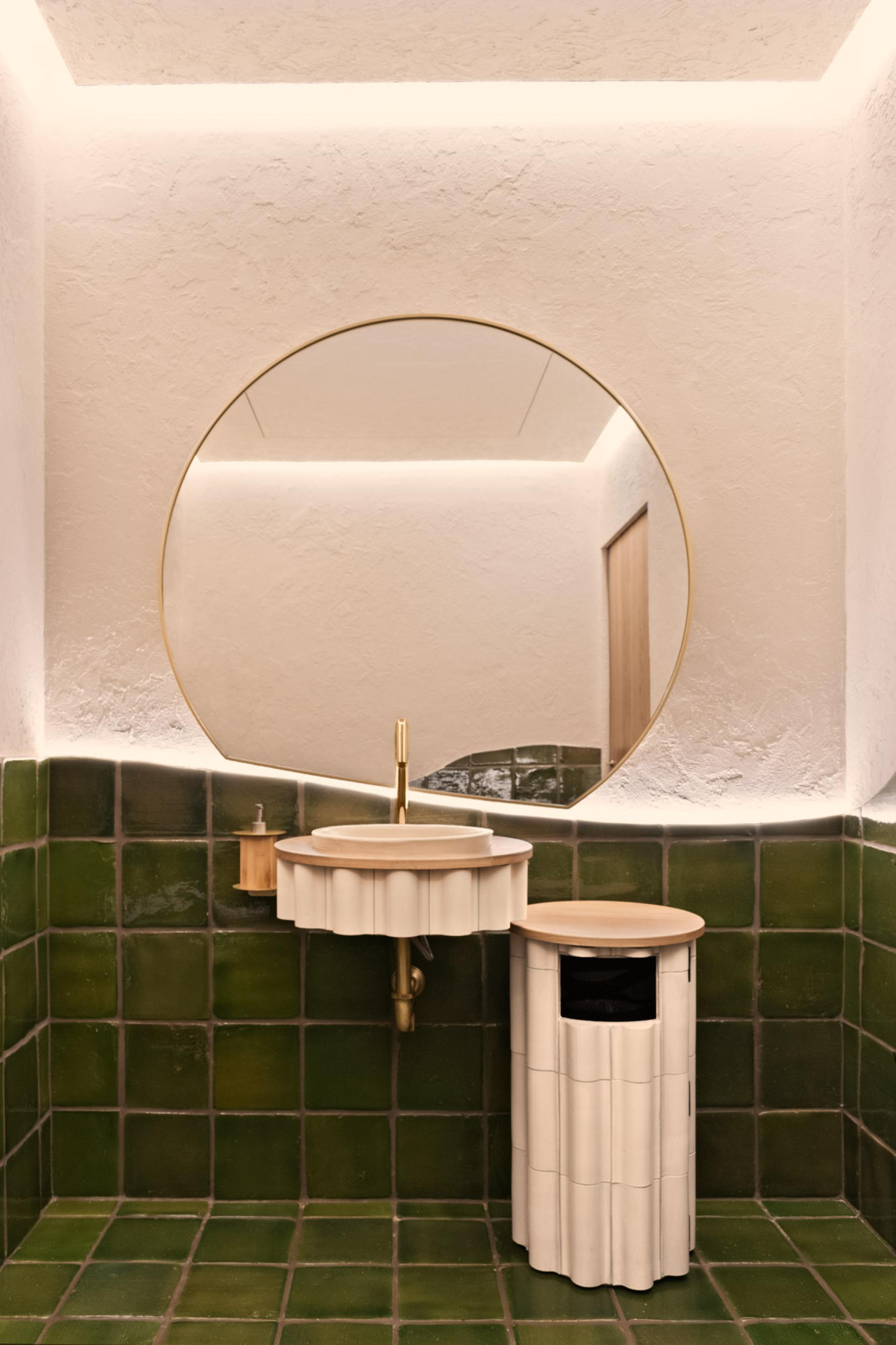 Modern restroom design featuring a round mirror above a sculptural sink and stylish green tiled walls.