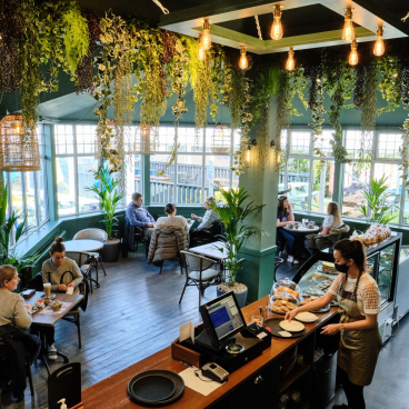 Cozy café interior featuring lush greenery, patrons enjoying food, and a barista preparing dishes in a biophilic design setting.