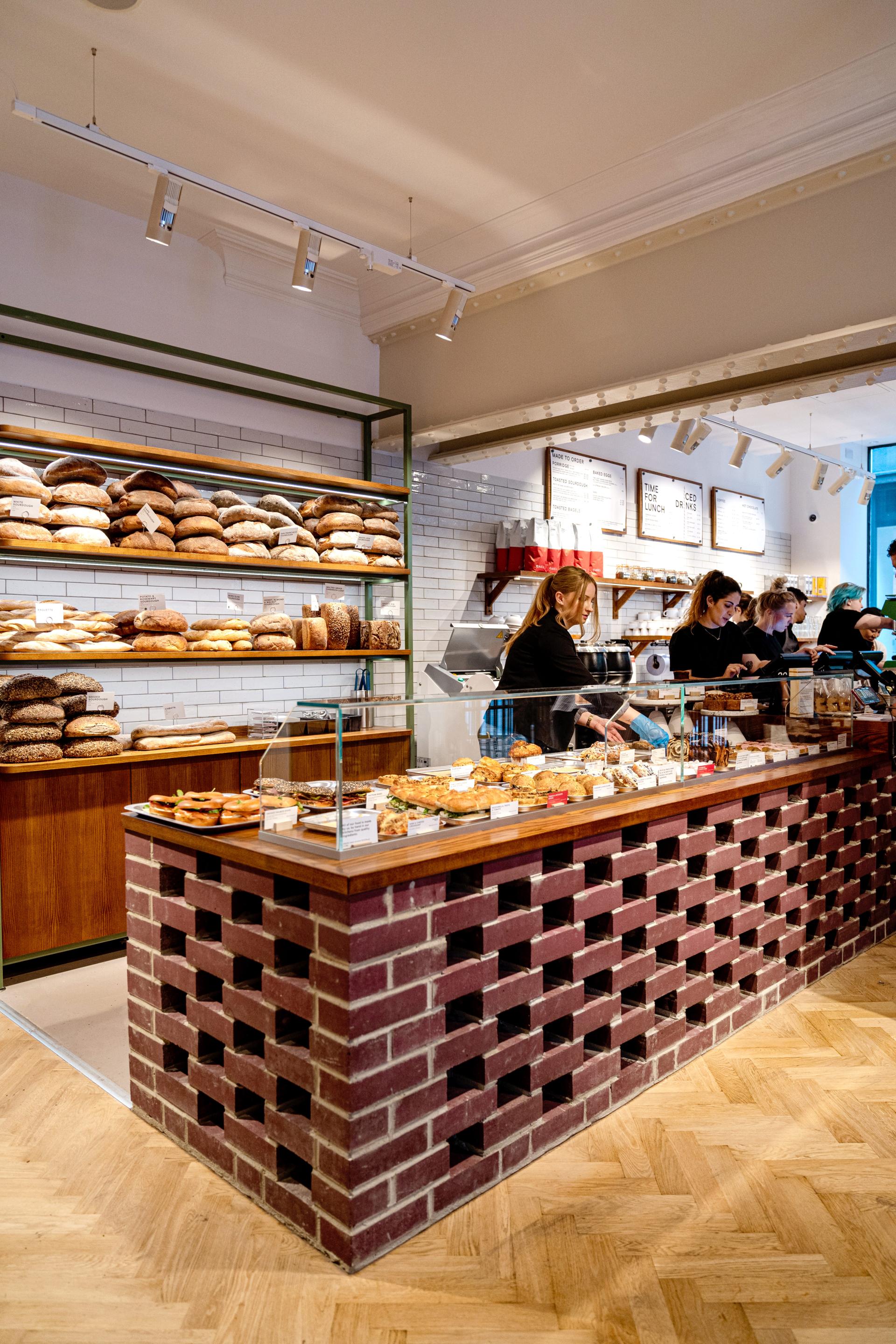 Modern bakery interior featuring a brick display counter, a variety of breads, and staff serving customers.