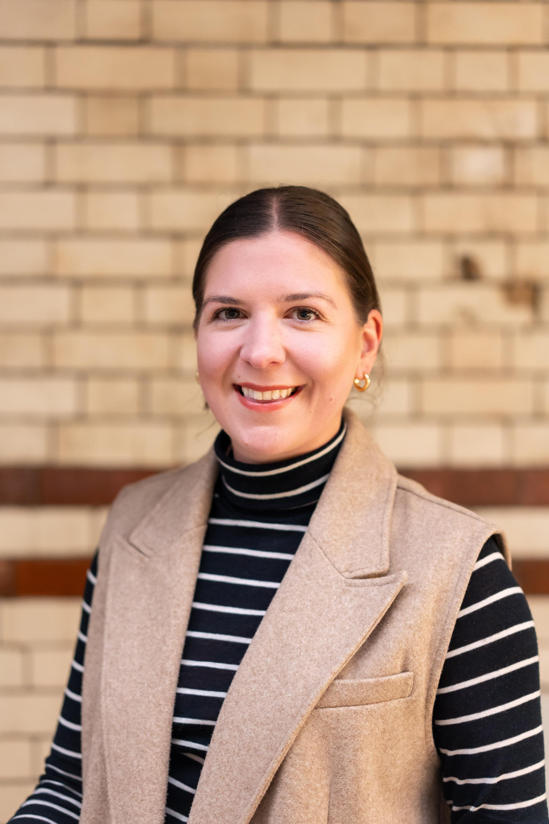 Smiling woman wearing a striped turtleneck and vest, against a textured brick wall, representing EDI culture discussion.