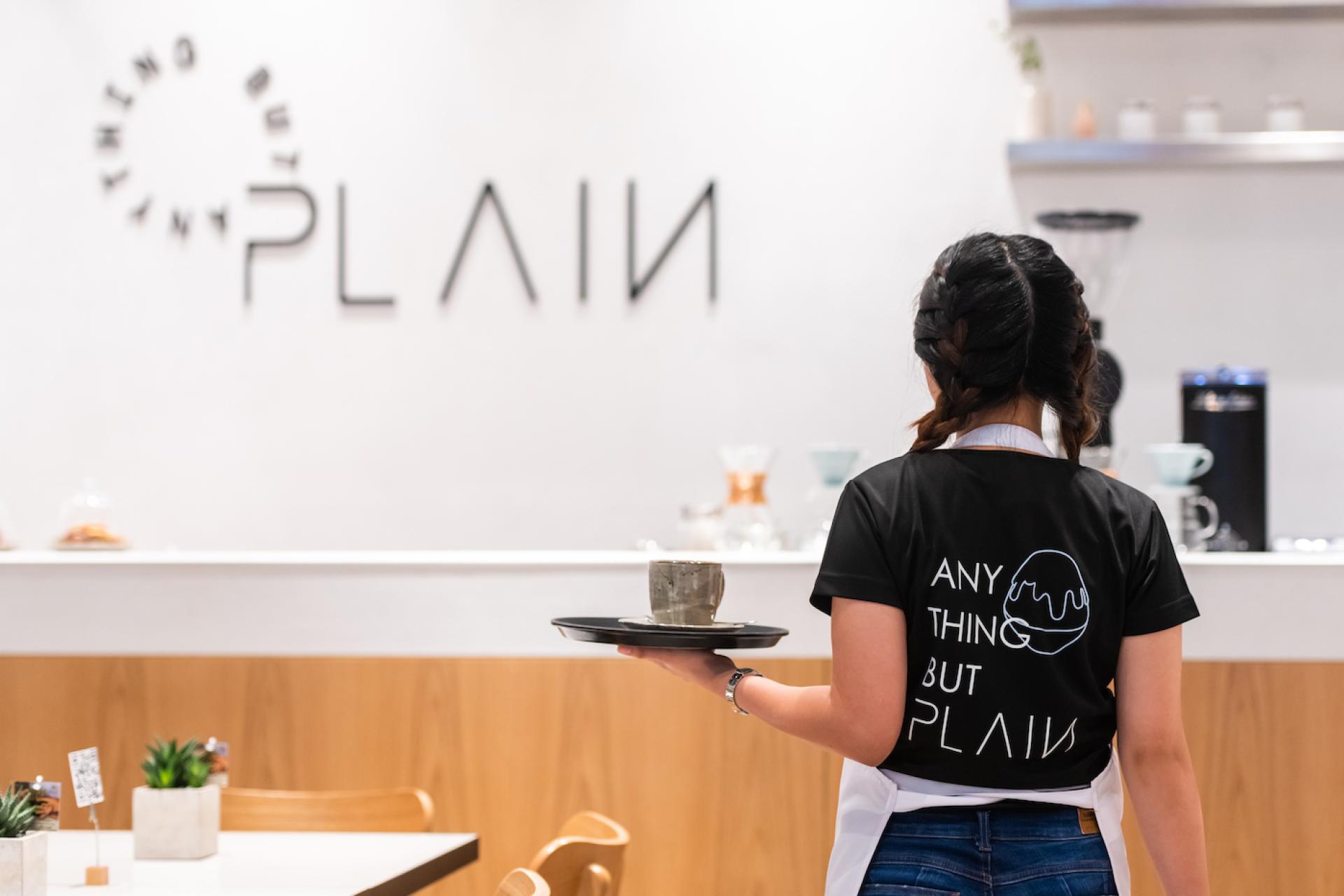Staff member in a modern cafe displaying handcrafted desserts, wearing a shirt featuring the brand name, "Anything but PLAIN."
