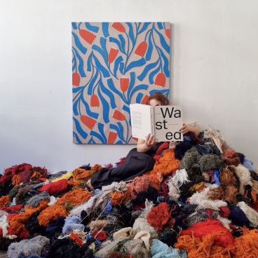 Sandra Junele sits amidst vibrant discarded textiles, reading a book on sustainable art and closed-loop practices.
