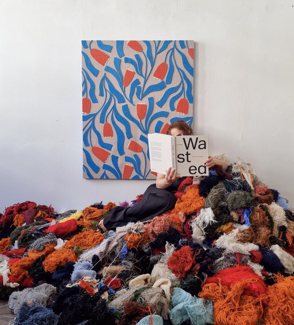 Sandra Junele sits amidst vibrant discarded textiles, reading a book on sustainable art and closed-loop practices.